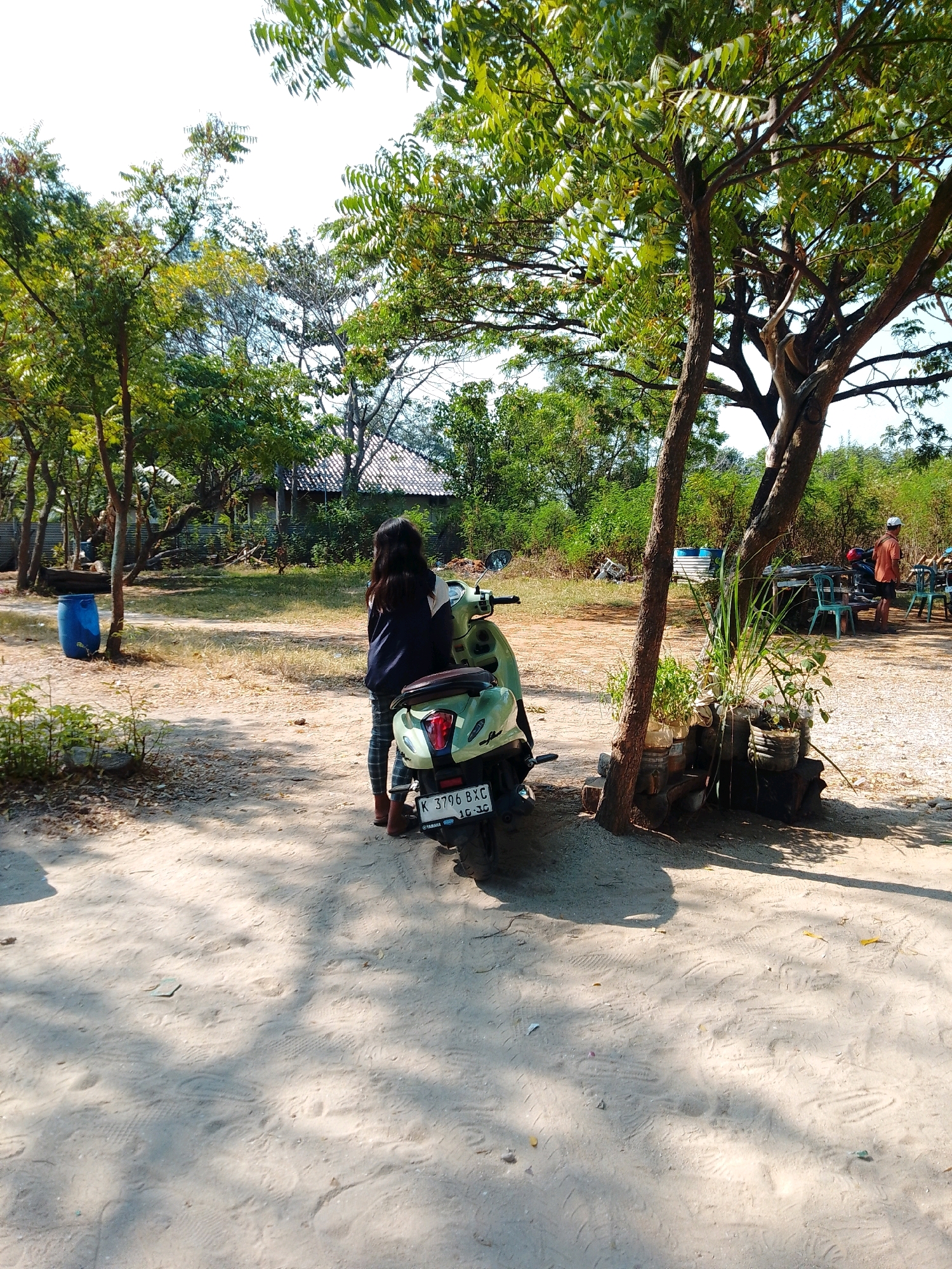 The image captures a sunlit outdoor scene, likely in Cikal, Indonesia. In the foreground, a light green scooter is parked on a sandy, dirt path. A person, viewed from behind, stands next to the scooter, wearing a dark jacket with white accents and plaid pants. The person's right foot is partially visible, wearing a brown boot.
The environment is characterized by several trees with green foliage casting shadows on the ground. In the background, a building with a tiled roof is visible amidst more trees and vegetation. To the right of the scooter, a group of plastic chairs and tables are set up under a tree, and a person in an orange shirt and shorts stands nearby. A blue barrel is positioned to the left of the scooter in the midground. The ground is covered with sand and dirt, with numerous footprints visible, suggesting it is a frequented area. The overall impression is of a casual, possibly rural or semi-rural, setting on a bright, clear day.