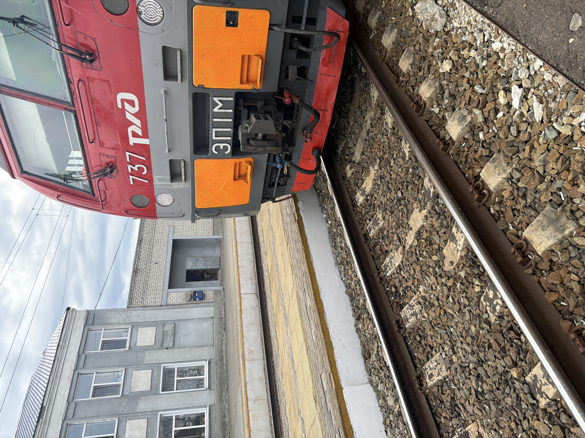 The image depicts a train station in Rtishchevo, Russia, with a red and grey train locomotive parked on the tracks in the foreground. The train has the number "737" and Russian text "РЖД" (RZD) on its front. To the left of the train, a two-story building with grey walls and several windows is visible. Adjacent to this building is a structure with a grey brick facade and an open doorway. The ground is covered with gravel, and a yellow-striped platform runs alongside the tracks. The sky is overcast with visible power lines. The scene is static, with no people or discernible actions.