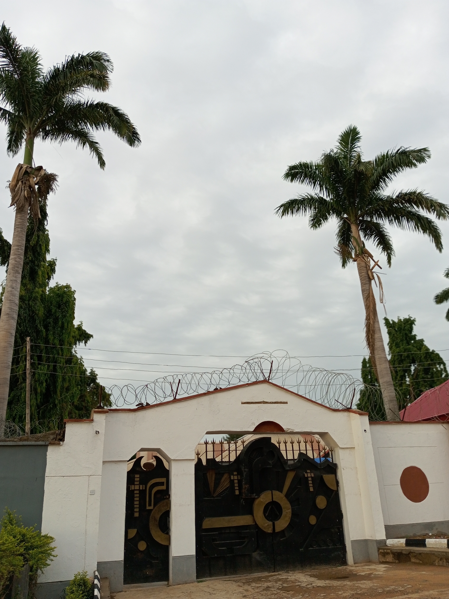 The image shows a closed black metal gate with intricate gold designs, flanked by white walls. Above the gate, there is a white arched structure with barbed wire coiled along its top. Two tall palm trees are visible on either side of the gate, with a line of green trees in the background. The sky is overcast with gray clouds. The scene appears to be an entrance to a property in Barden Yana, Nigeria.