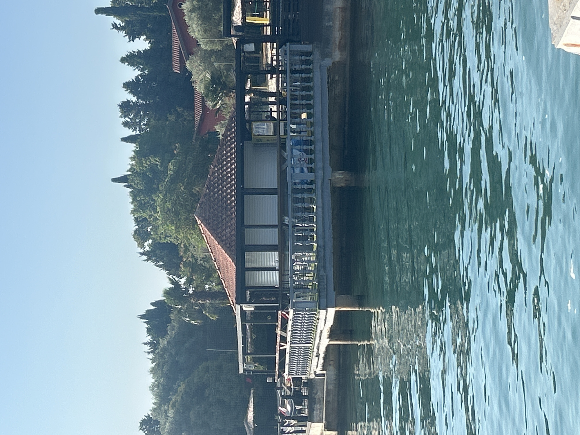 The image depicts a waterfront scene, likely in Monterol, Croatia, under a clear blue sky. In the foreground, the turquoise water is rippled, reflecting the light. A pier or walkway juts out into the water, supported by concrete pillars. On this structure, there is a small building with a terracotta-tiled roof, resembling a kiosk or small cafe. The building has closed shutters or blinds on its windows. The pier is lined with a decorative balustrade. In the background, lush green trees and vegetation form a dense backdrop, with some cypress trees visible. Beyond the trees, red-tiled roofs of other structures can be seen. The overall impression is one of a calm, sunny day at a coastal location. No people are clearly visible in the image.