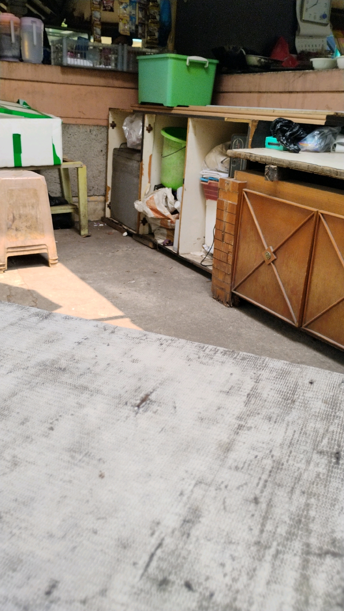 The image depicts a cluttered outdoor stall or storage area. In the foreground, a textured grey mat covers the ground, with a distinct diagonal shadow cast across it from an unseen object. To the left, a light brown plastic stool is visible, partially shadowed. Behind the stool, a white and green box rests on a yellow-legged stand.
The mid-ground reveals an open storage unit with wooden shelves. On the top shelf, a bright green plastic bin with a lid sits prominently. Below this, an open compartment reveals a green bucket, plastic bags, and assorted items including what appears to be fabric or paper. To the right of this storage unit, a wooden gate with a diamond pattern is partially visible, supported by brick pillars. The environment appears to be an outdoor, possibly commercial or residential, space with a generally unkempt appearance. There are no people or animals visible in the frame. The location is identified as Central Jakarta, Indonesia.