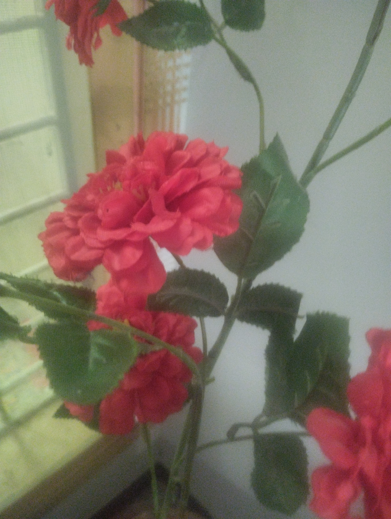 A close-up, vertically oriented, medium shot captures an arrangement of bright red, artificial flowers and green leaves against a pale, neutral background. The focus is on the textured petals of the flowers, which appear to be roses or peonies, with many layers of tightly packed petals. The leaves are a deep green, some cupping the flowers, others more spread out. The stems are thin and green, with a few small buds or developing leaves visible. The lighting is soft and diffuse, casting no harsh shadows. The overall impression is one of artificial beauty and stillness, with the flowers appearing to be part of a decorative display. The background is slightly out of focus, with hints of muted yellow and white that suggest an indoor setting, possibly a room with a window. The image quality is somewhat grainy, indicating it may have been taken with a low-resolution camera or in low light conditions.