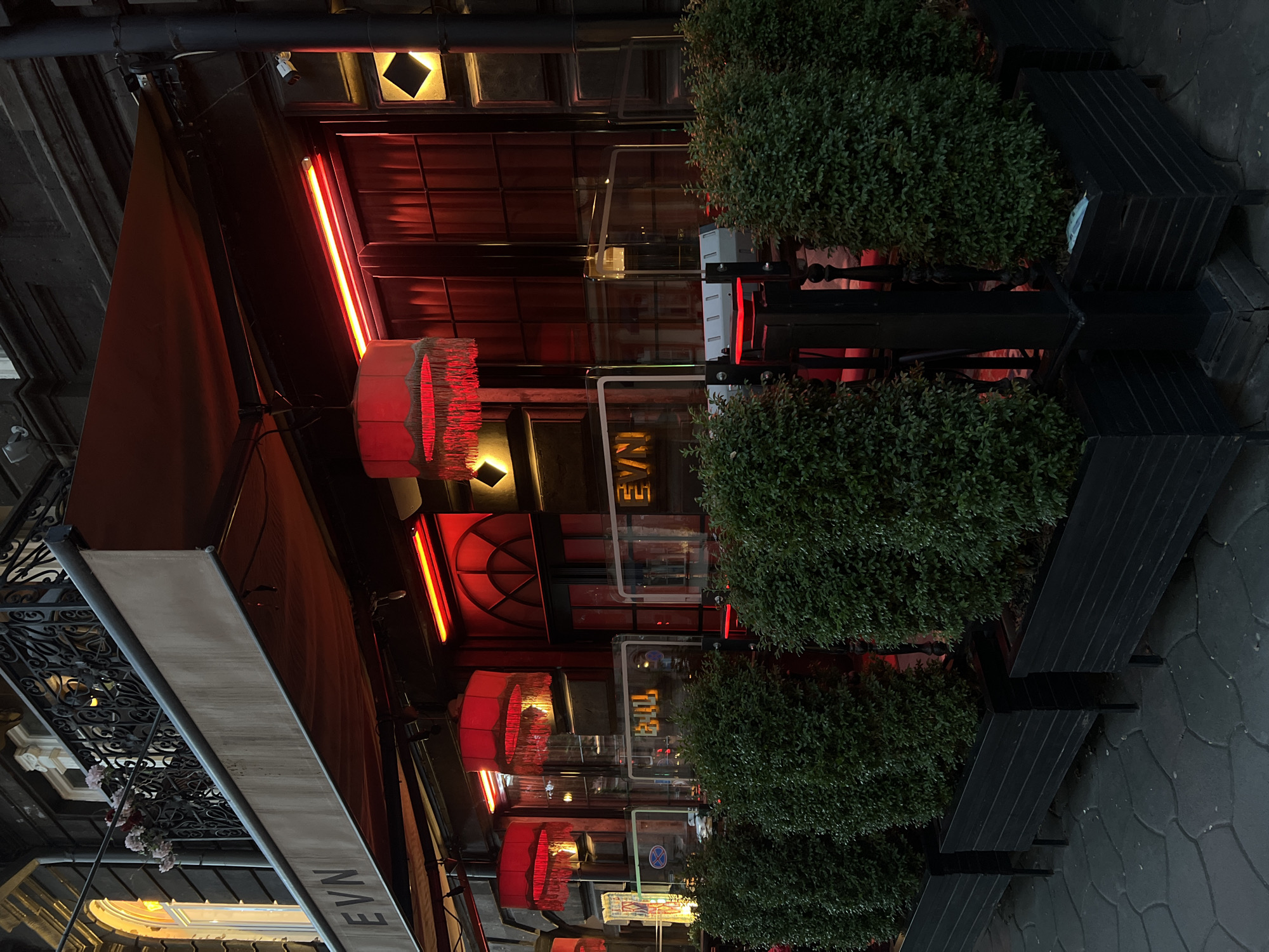An exterior shot of the storefront of a business in an unknown location at dusk. The façade is adorned with an awning, red neon lights, and cylindrical red hanging lamps. The business name, EVN, is seen on the awning. The entrance has a glass door with illuminated signage. Three potted plants flank the entrance, and black benches provide seating. The sidewalk is paved with a patterned design. The lighting creates a warm ambiance. No people are visible.