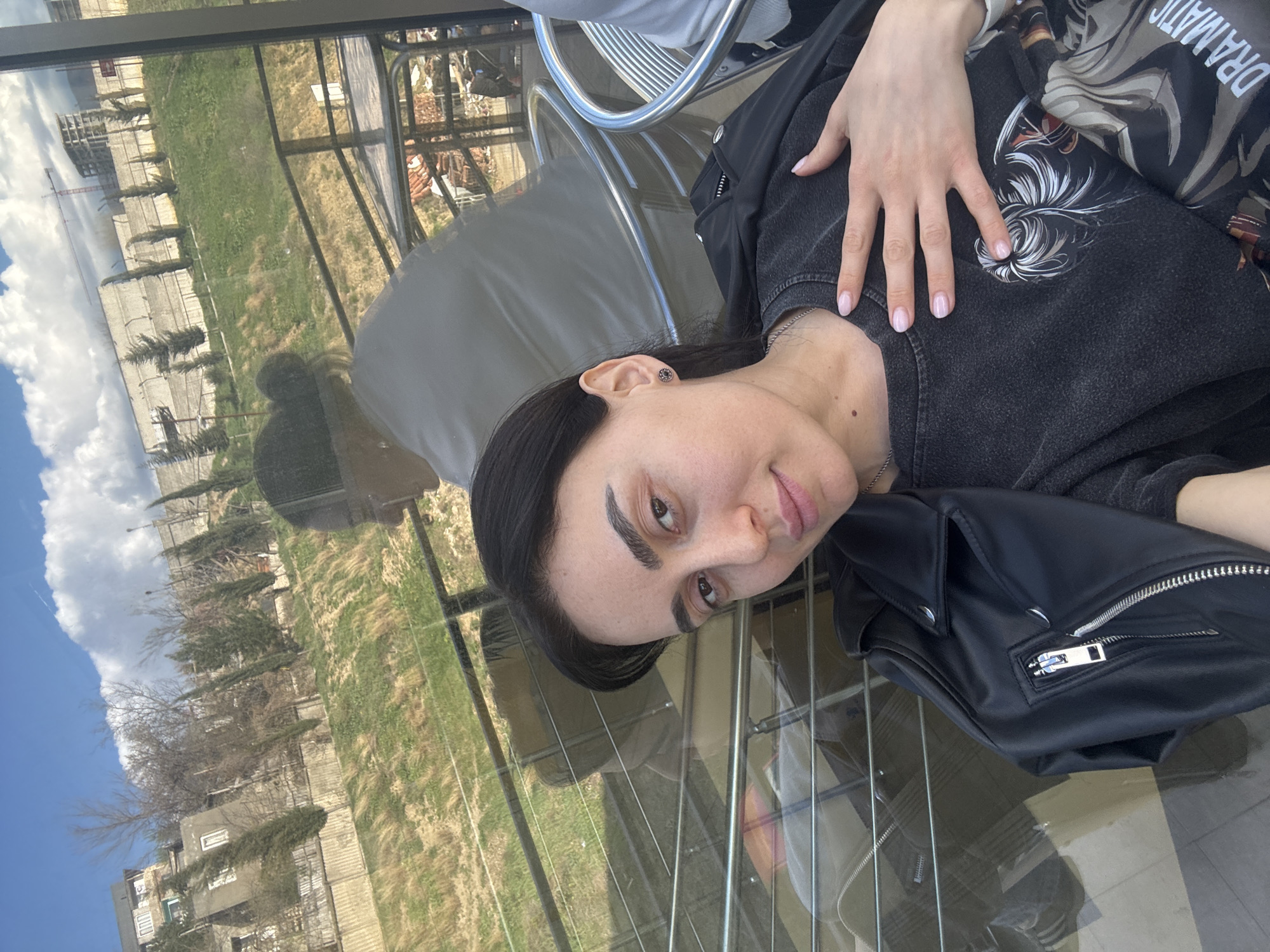 A woman with dark hair is seated at a table outdoors. She wears a dark-colored top with a design, a black jacket, and a necklace. Her left hand rests on her upper chest area. The table has a glass top reflecting the outside environment, which includes buildings, trees, and a cloudy sky. The location is likely Tbilisi, Georgia, given the architectural style of the buildings and the style of vegetation. The seating arrangement appears to be in a cafe or restaurant setting.