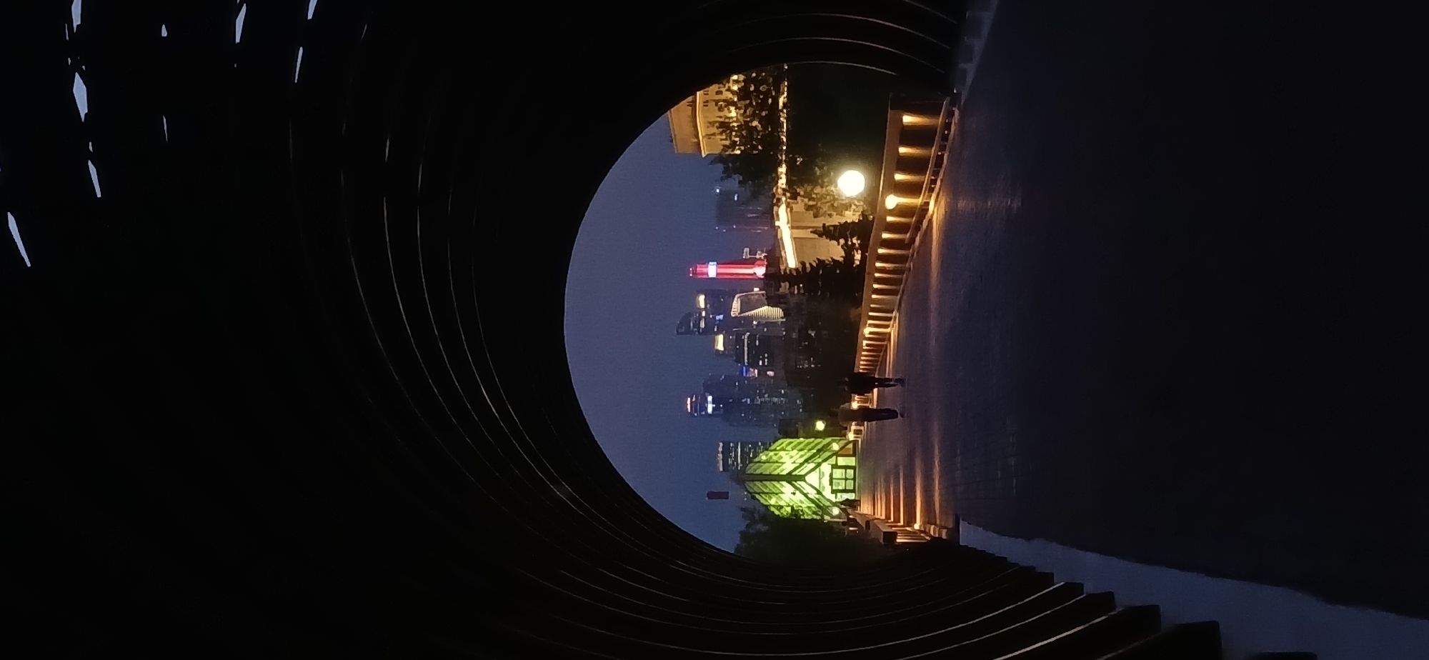 A photograph presents a night-time urban landscape viewed through a circular architectural structure, likely a tunnel or frame. The location features a cityscape with illuminated skyscrapers, including one with a distinctive red top. A pathway is visible, lined with lights and what appears to be a building with green lighting at the bottom. Two individuals are present, walking along the pathway. The scene is generally dark, with the city lights serving as primary sources of illumination. The photograph has been rotated.