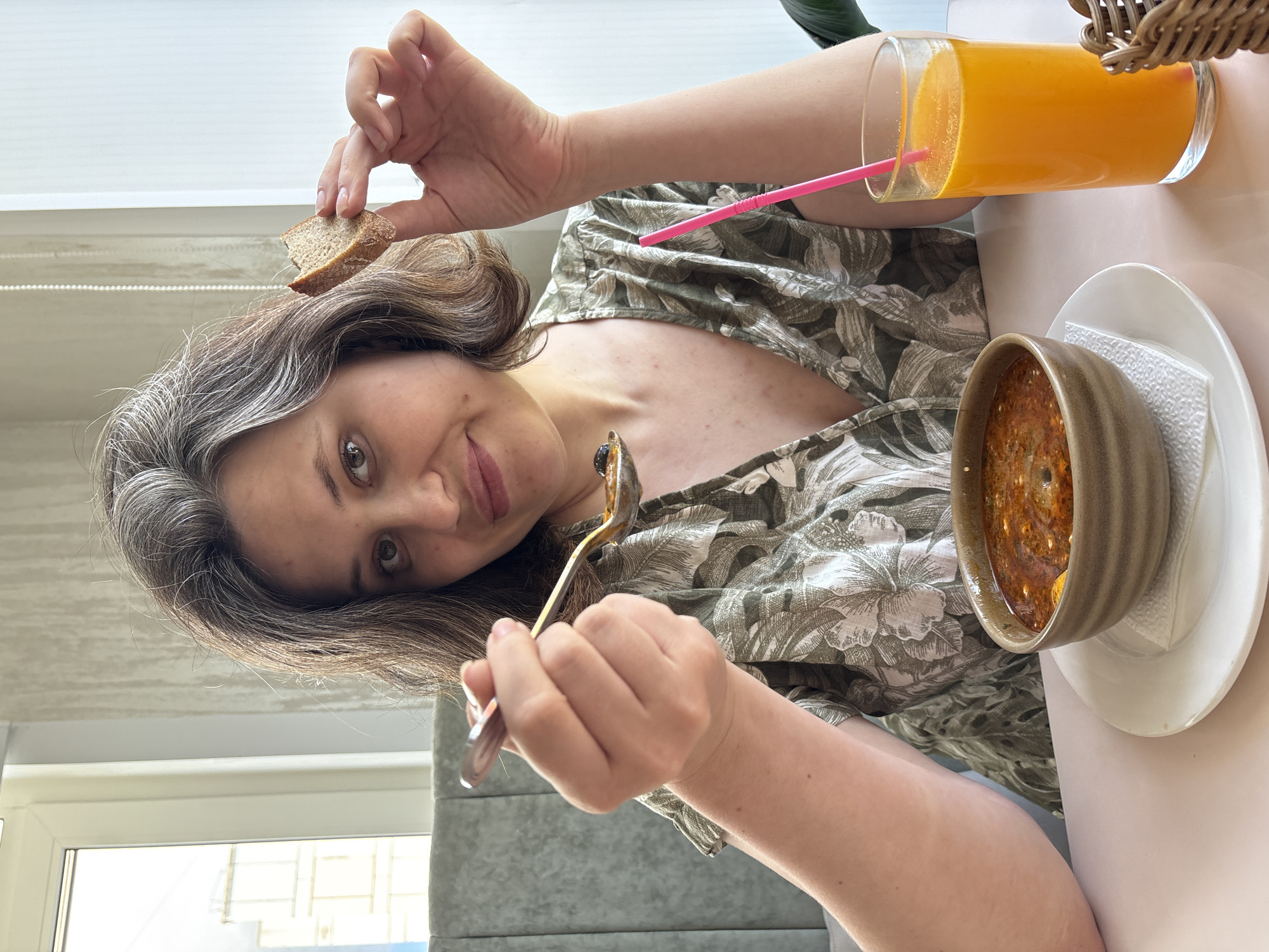 A woman sits at a table in what appears to be a restaurant or cafe, enjoying a meal. She is holding a small piece of bread in one hand and a spoonful of soup in the other, looking towards the camera with a slight smile. On the table in front of her is a bowl of soup on a white plate, and a tall glass of orange juice with a pink straw. The setting includes a window, a portion of a woven basket, and a neutral-toned wall. There is nothing to suggest the location as the city of Chaikovsky, Russia, is identifiable from the image.