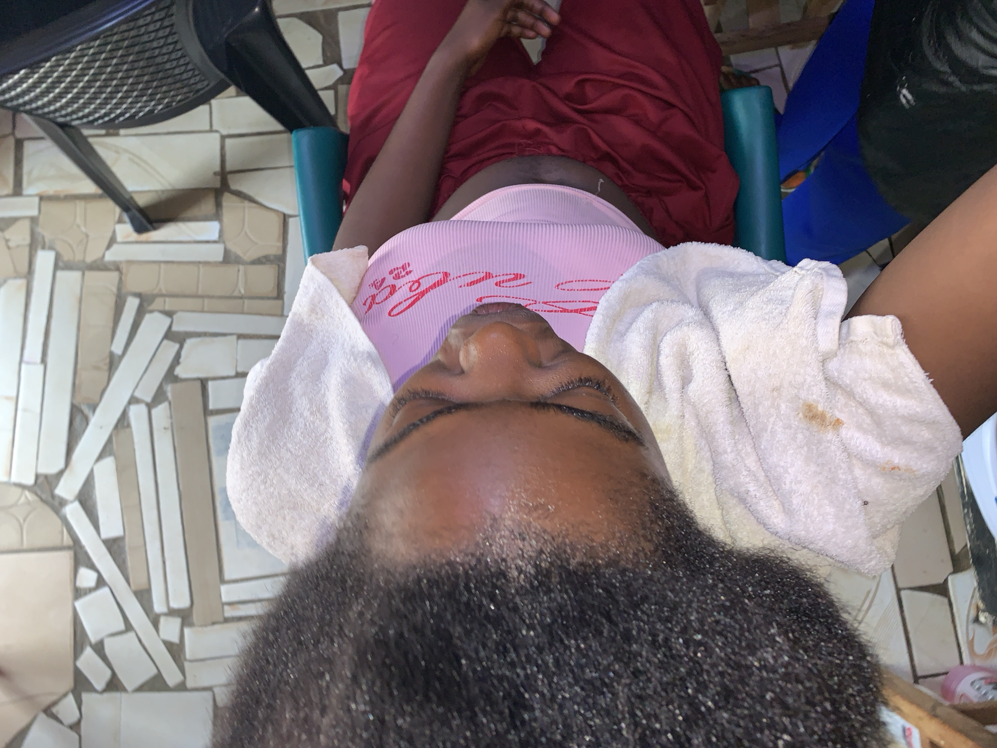 A woman is lying in a chair with her head tilted back, looking up at the camera. She is wearing a pink tank top with red writing and red pants. A white towel is wrapped around her head. Her hair is dark and curly. The chair is teal and black. The floor is made of light-colored tiles arranged in a random pattern. The scene is located in the city of Rafin Par, Nigeria.