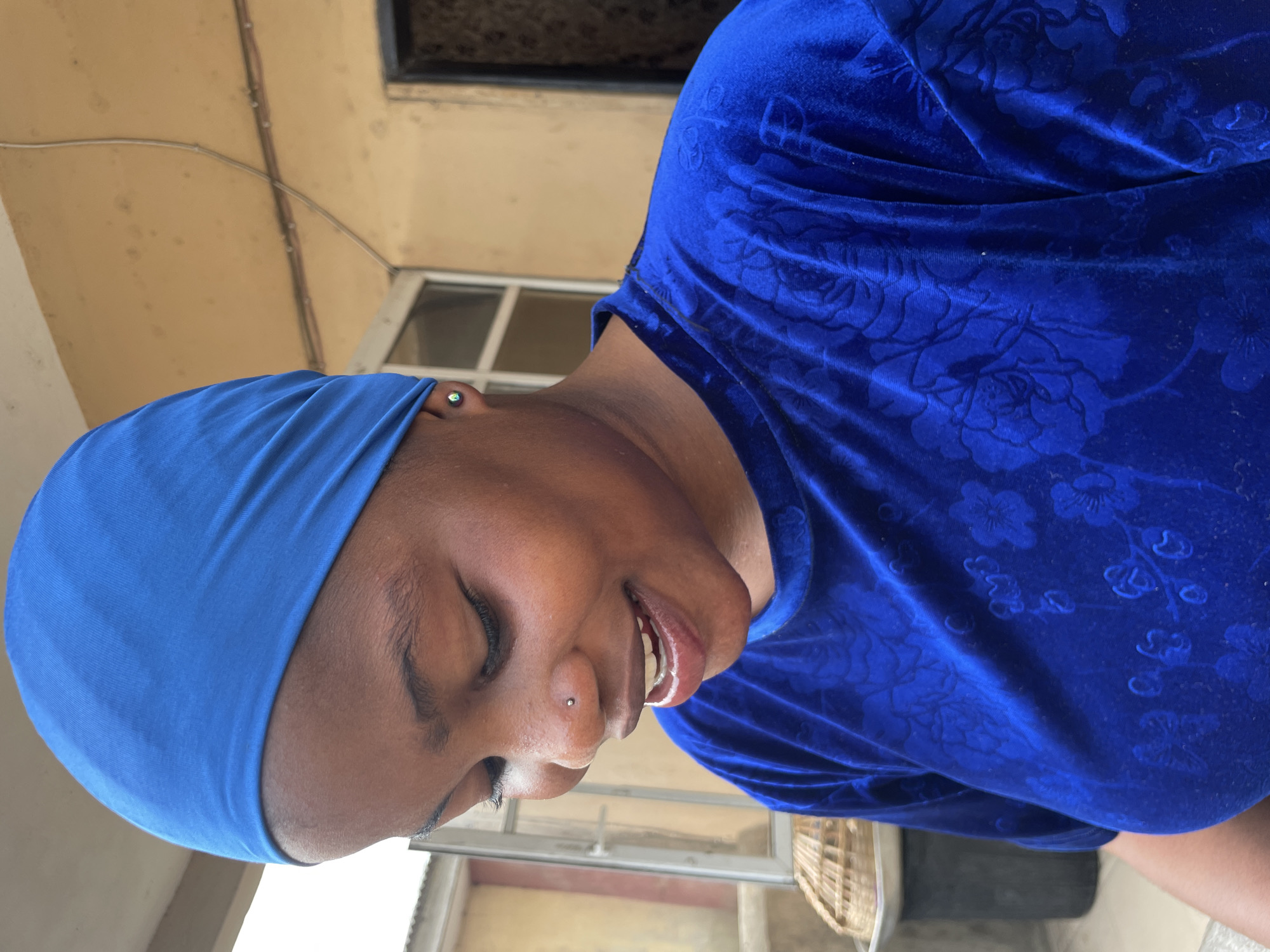 A medium shot of a woman with dark skin wearing a blue headscarf and a blue velvet top with a floral pattern. She has her eyes closed and is smiling. Her ear is pierced with an earring and her nose is pierced with a stud. The background includes a building exterior with a window and a tan wall. There is a wicker basket in the lower right corner. The location is Badagry, Nigeria.