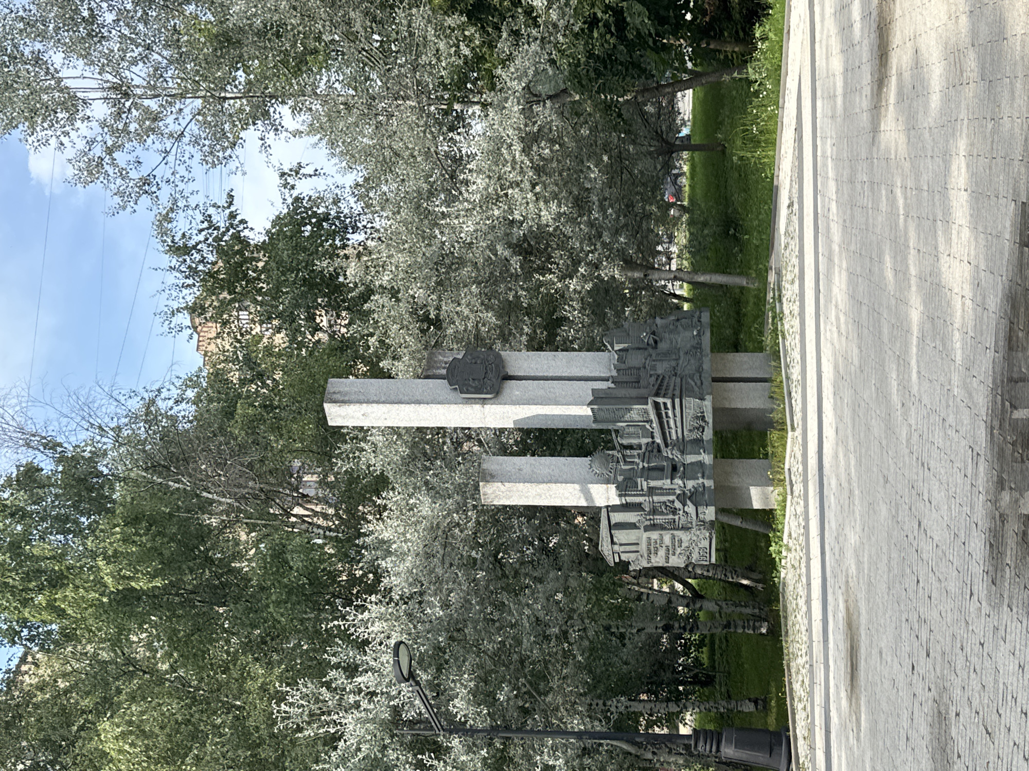 The image captures a monument in Zelenograd, Russia, located in a paved public square surrounded by trees and a building in the background. The monument itself is made of granite and features two tall pillars with a relief sculpture at the base. The relief depicts a cityscape with buildings, a sun, and what appears to be a train. Inscriptions are visible on the monument, including "1958" and Cyrillic text. A lamppost stands to the left of the monument. The ground is paved with light-colored stones, and there is grass and foliage around the monument. The sky is blue with some clouds. No people are visible in the frame.