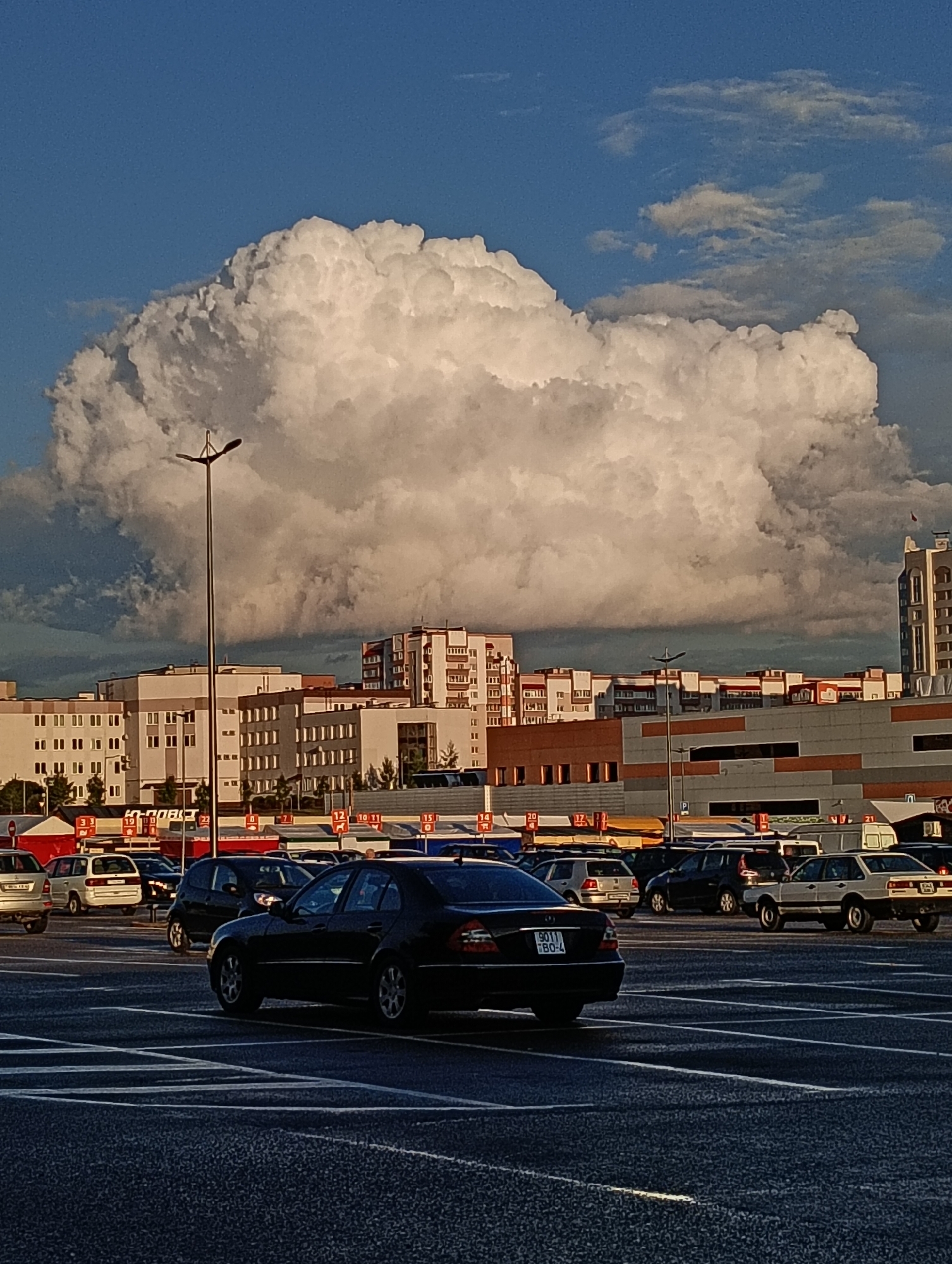 In the city of Hrodna, Belarus, a vast, bulbous cumulonimbus cloud dominates the sky over a wide, asphalt parking lot filled with vehicles. The cloud, illuminated by the sun, displays a range of white and grey tones, creating a dramatic contrast against the clear blue sky to its left. In the mid-ground, a line of low-rise and mid-rise buildings, typical of urban architecture, runs horizontally across the scene. Some buildings are rendered in pale colors with visible windows, while others feature more modern facades with horizontal bands of darker material. A tall, dark lamppost with two lamps stands prominently in the foreground on the left, silhouetted against the massive cloud. The parking lot itself is marked with white lines indicating parking spaces and driving lanes. Several cars are visible, including a black sedan in the foreground, and various other vehicles like SUVs and smaller cars parked and driving within the lot. The overall impression is a typical urban landscape under a striking, cloudy sky, likely captured during daylight hours.