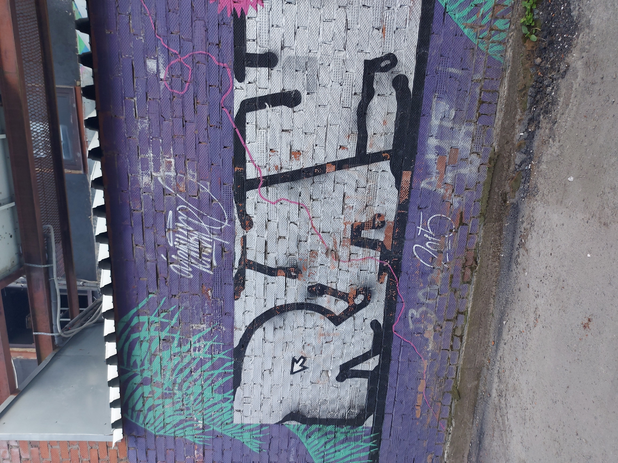 A vertical shot shows a brick wall, painted purple with graffiti, located in Izhevsk, Russia. The graffiti consists of a large, stylized white word outlined in black, with the letters arranged vertically. A smaller, cursive script in white is above this, and "2015" is written to the right of the large word. Pink lines are woven among the graffiti. The lower portion of the wall contains green palm-like fronds. The building on the left of the image has steel beams and a corrugated awning. The edge of a gray road or pavement is seen on the right.