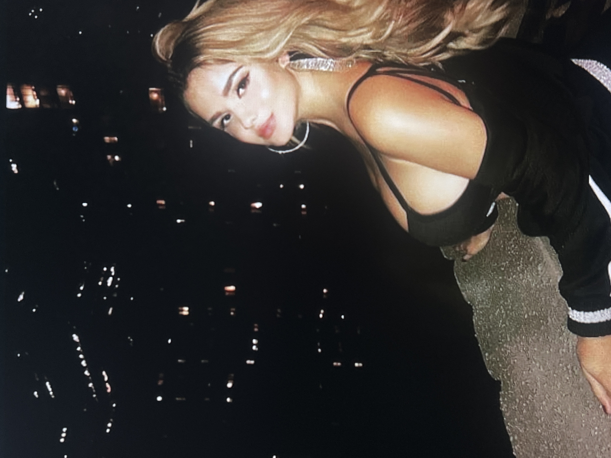 The image shows a woman with long, blonde hair and large hoop earrings. She is wearing a black top with thin straps and a black jacket with white stripes on the sleeves. She is leaning forward, with her head tilted down, and her eyes are looking towards the camera. The background is dark and appears to be a cityscape at night, with many small lights visible. There is a rough, textured surface in the foreground, possibly a stone ledge or wall. The overall mood of the image is somewhat dramatic and alluring, with the lighting and the woman's pose creating a sense of mystery. The time of day is clearly night. There is no visible text.