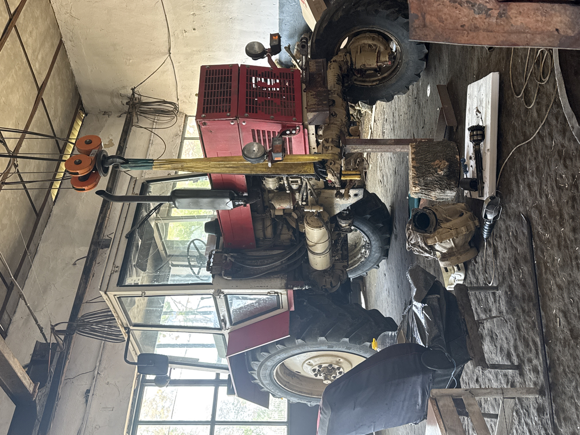 The image captures the interior of a garage or workshop in Sarnevo, Bulgaria, with a large, older model red and white tractor as the central subject. The tractor appears to be undergoing maintenance or repair, as its front end is elevated. A yellow strap is attached to the front of the tractor, connected to an industrial lifting system featuring orange pulleys with "2t" visible, indicating a 2-ton capacity. A wooden log is placed beneath the front axle, seemingly providing additional support.
The tractor has a cab with glass windows, a prominent exhaust pipe, and visible engine components, hoses, and wires. Its rear wheels are large and treaded, with "DUNLOP" and tire size information (e.g., "6.50-20") partially legible on the sidewall. The front of the tractor features red grated covers and headlights.
The surrounding workshop environment is utilitarian, with concrete or plastered walls showing signs of age and wear. Exposed wooden beams are part of the ceiling structure. Natural light streams in through several windows, suggesting it is daytime. Various workshop items are scattered around: tangled electrical cords or wires hang from a beam, a white board with an attached light fixture or tool lies on the dirt floor, alongside a black tarp or cover, a ladder, and what appears to be a detached transmission or engine part. The overall setting indicates an active, functional workspace. No people are visible in the scene.