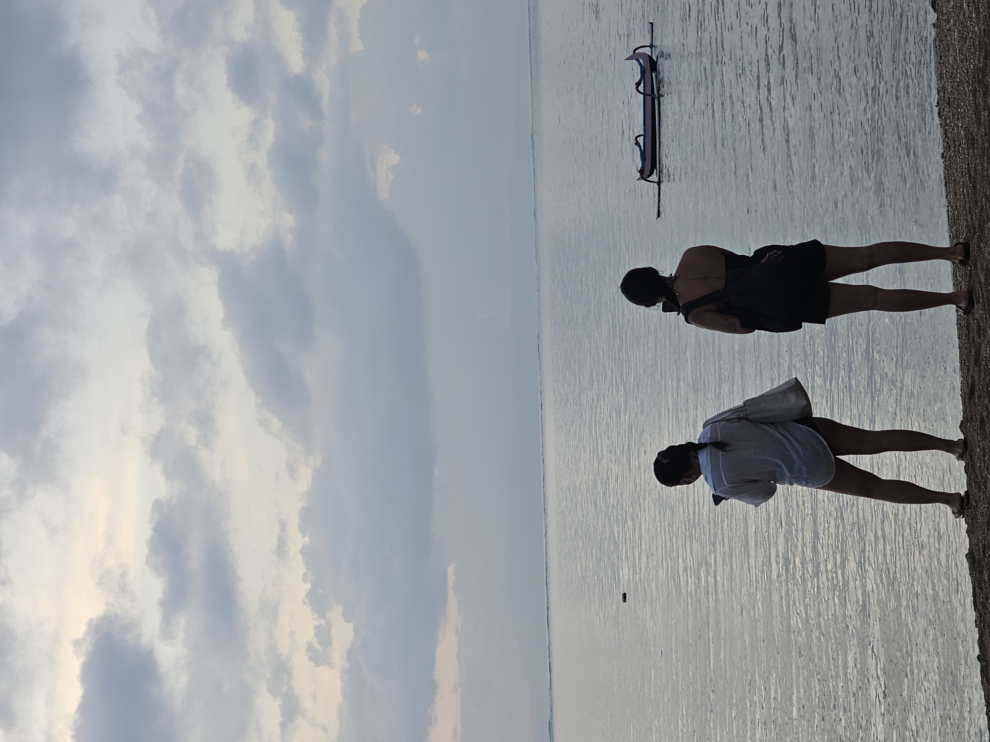 The image displays two individuals standing near the water's edge. The person on the left is wearing a black dress and has their arms crossed. The person on the right is wearing a white shirt and carrying a bag over their shoulder. In the background, a purple inflatable object is visible on the water, possibly a tube or raft. The scene appears to be a beach or shoreline, with the water extending to the horizon. The sky is filled with clouds, suggesting it might be overcast or nearing sunset or sunrise. The overall mood is calm and serene. The image appears to be taken in Denpasar, Indonesia.