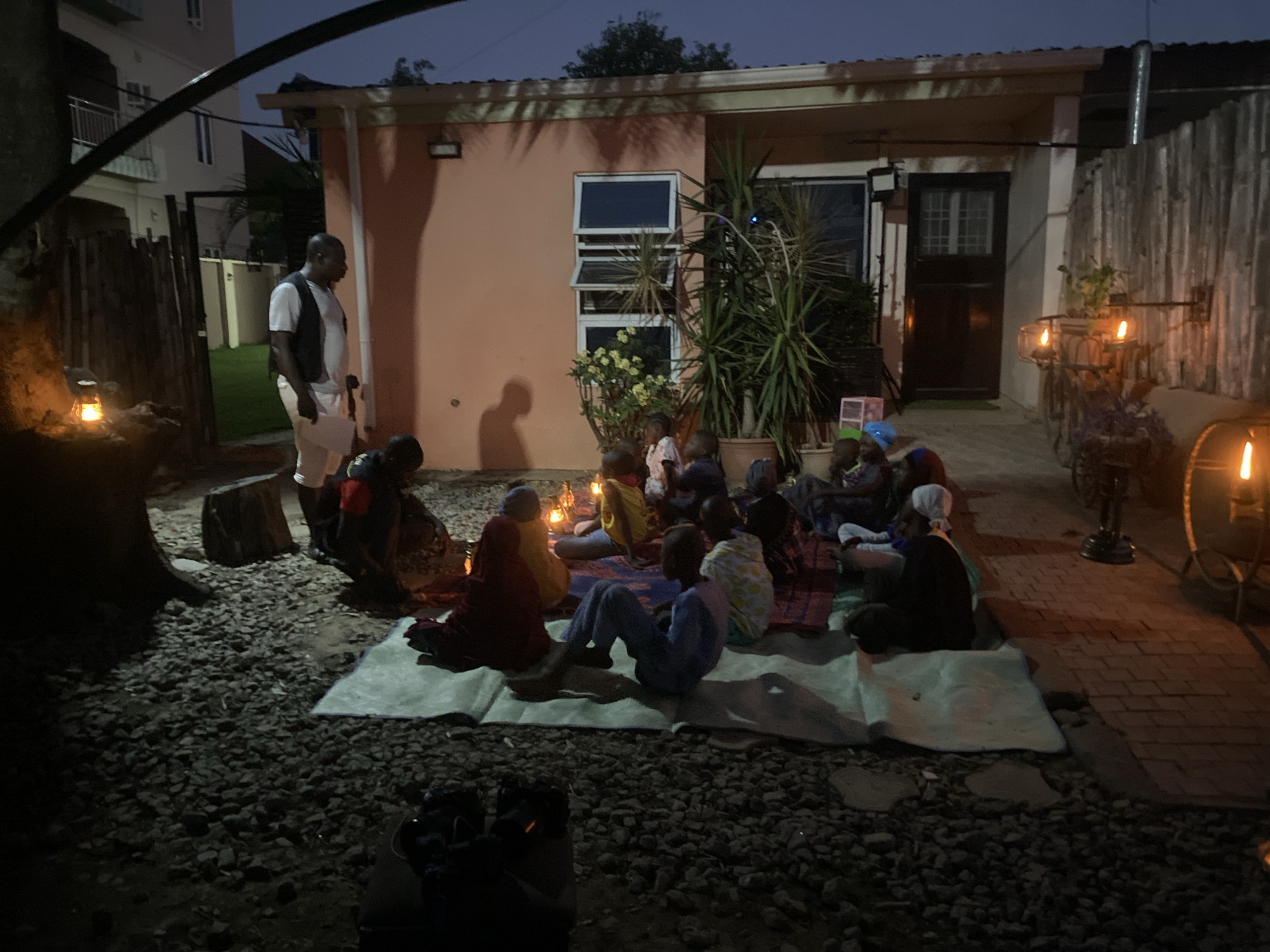 A group of people, mostly children, are gathered outside at dusk. They are seated on blankets and mats on a gravel surface. A man stands nearby, observing the group. A building is in the background, with windows and plants visible. Light is provided by several lanterns placed around the scene. There's no indication of the location within Barden Yana, Nigeria.