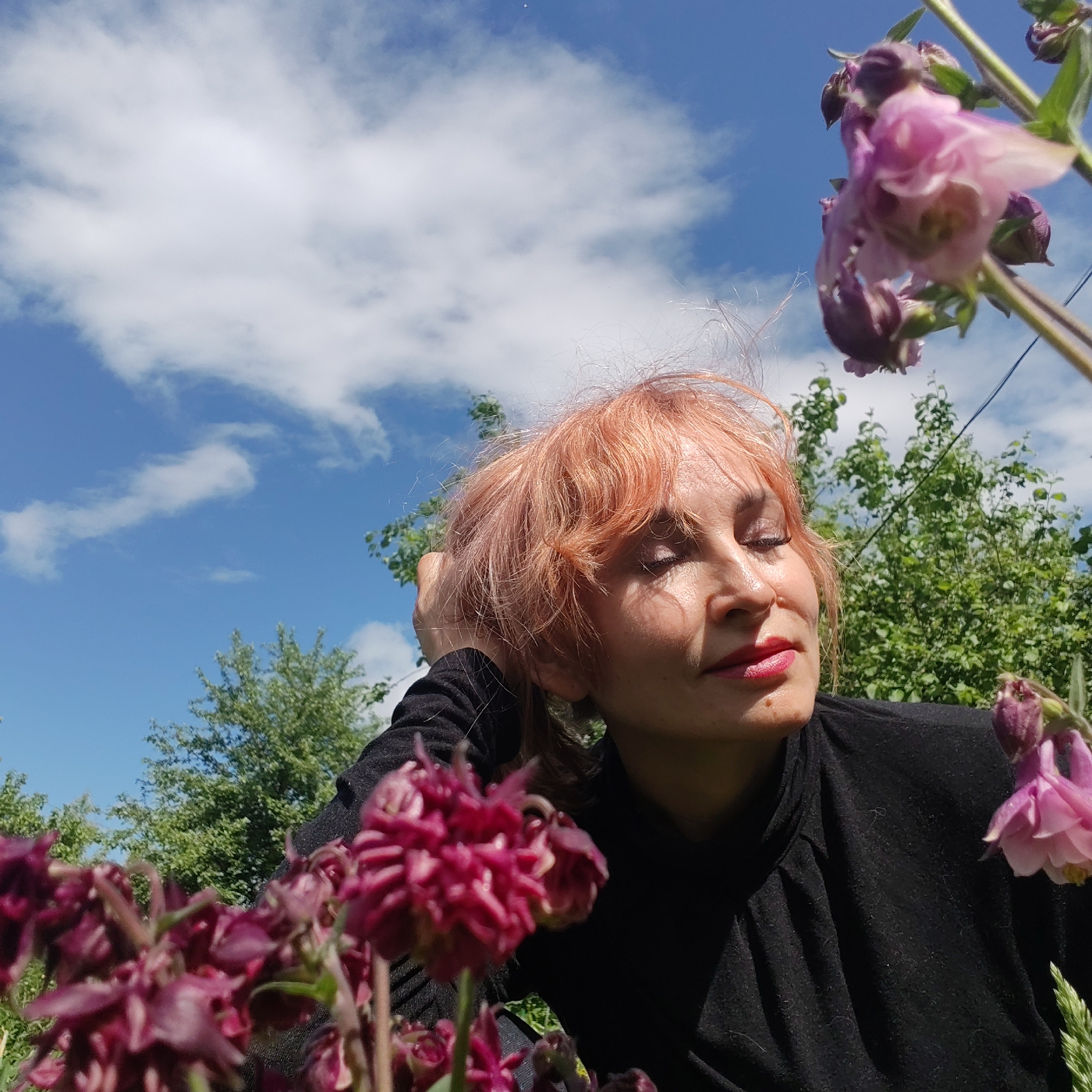 The image shows a woman with short, light-orange hair and red lipstick in an outdoor setting. She has her eyes closed and is angled upwards towards a blue sky with white clouds. She is wearing a black long-sleeved shirt. The foreground is filled with purple and pink flowers, while green trees are visible in the background. The specific location within the city of Chaikovsky, Russia is not discernible from the image.