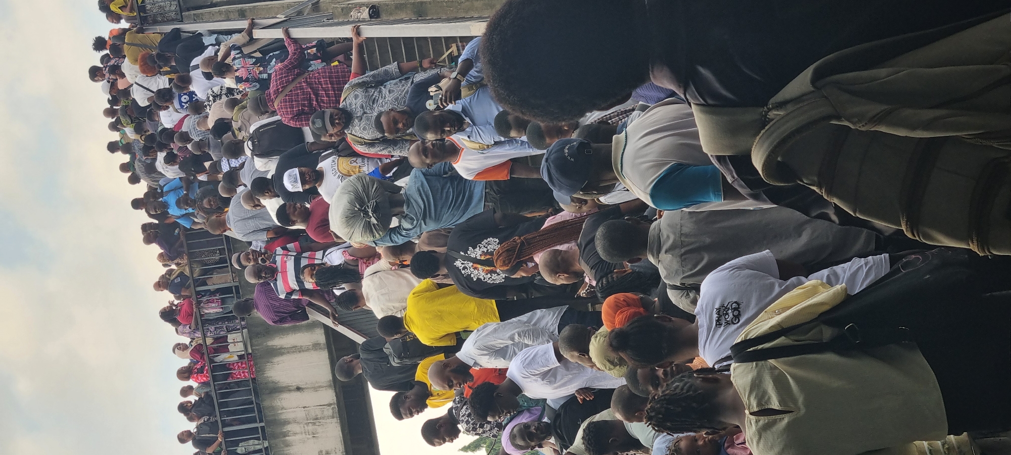 This is a photograph taken in Ikeja, Nigeria, depicting a large crowd of people ascending a concrete pedestrian overpass. The overpass has a metal railing along the left side, which is lined with people. The crowd is densely packed, with individuals of various ages and genders, predominantly appearing to be adults. Many are dressed in casual clothing, including t-shirts, shirts, and trousers. Several individuals are carrying bags, including a person in a blue denim shirt carrying a large, light-colored bundle on their head. Some people are looking down, others ahead or at their phones. The environment appears to be an urban setting, with the sky visible in the upper portion of the image, indicating an outdoor scene. The lighting suggests it is daytime.