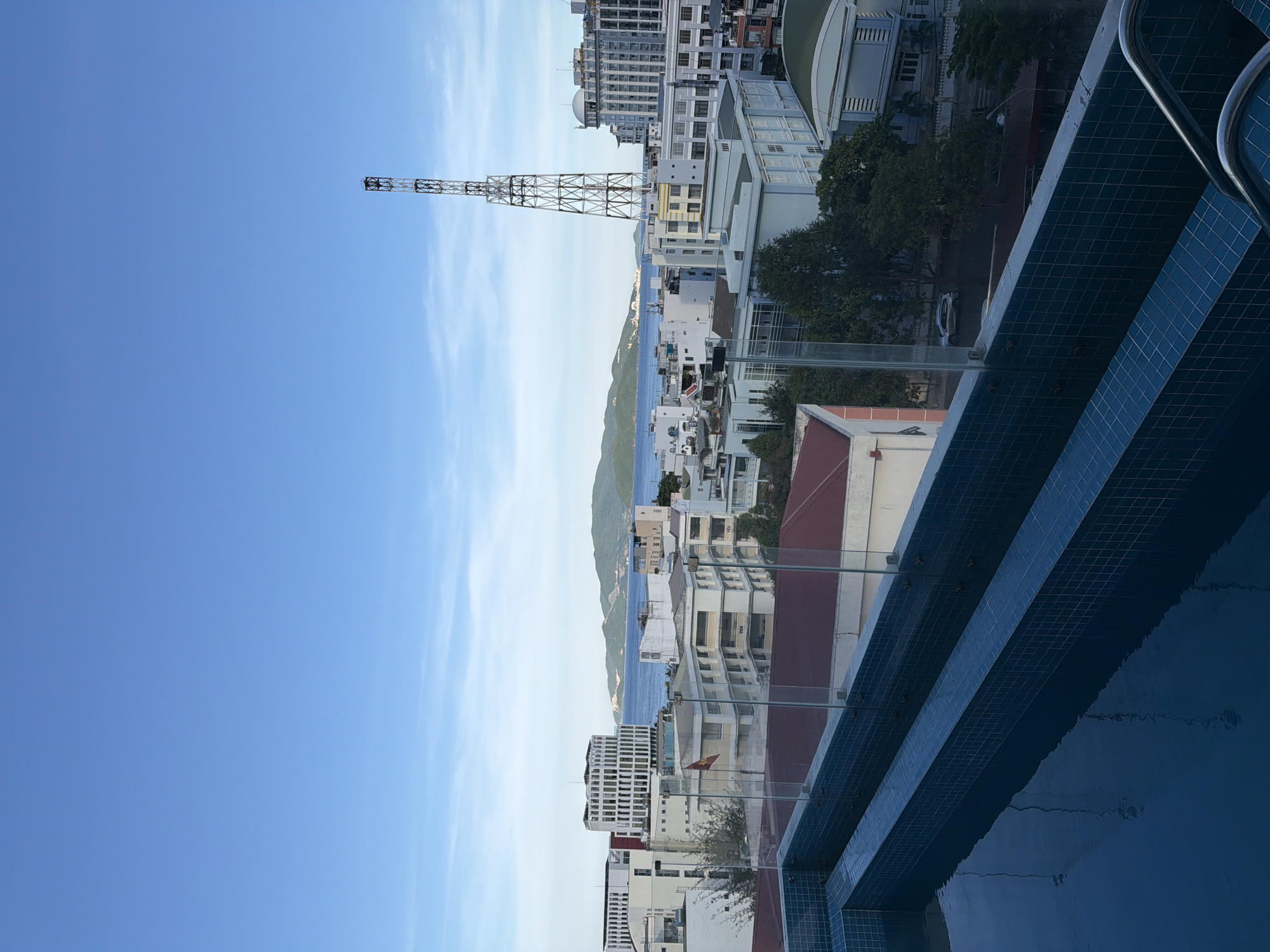A panoramic view captures the cityscape of Nha Trang Ward, Vietnam, under a clear blue sky. In the foreground, a partially visible swimming pool with blue tiled edges and glass railings suggests a rooftop setting. Beyond the pool, the city unfolds with various buildings, ranging from multi-story structures with modern architecture to older residential and commercial buildings with red tiled roofs. A prominent lattice telecommunications tower stands tall amidst the urban landscape, reaching towards the sky. In the distance, the turquoise waters of the sea meet a lush green island, creating a scenic backdrop to the city. The scene appears peaceful and serene, with no visible human activity. The overall impression is one of a tranquil morning or late afternoon, offering a glimpse of coastal urban life in Vietnam.