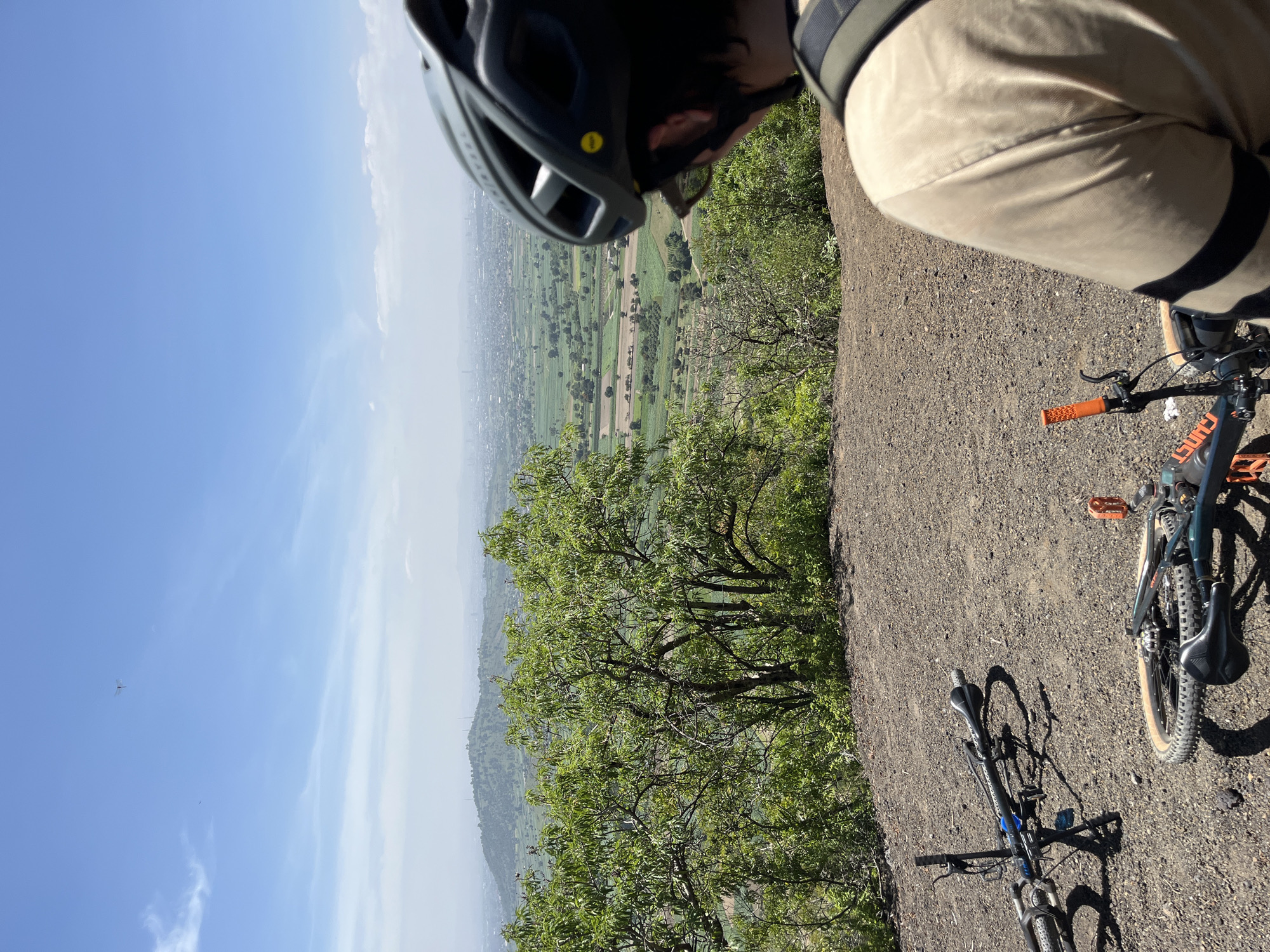 This image depicts a scenic overlook from a hillside, likely in the vicinity of San Miguel Papaxtla, Mexico. In the foreground, two mountain bikes are resting on a gravelly surface. One bike is upright and appears to be secured with a kickstand, while the other is lying on its side. A person wearing a beige shirt and a dark helmet is visible from the shoulders up, facing away from the camera and looking out at the expansive landscape. The environment features lush green trees and vegetation in the mid-ground, with a hazy view of a city or town spread across the valley in the background. The sky is bright blue with scattered clouds, and a tiny, unidentified flying object can be seen in the upper portion of the frame. The overall scene suggests a pause during a bicycle ride, taking in the panoramic view of the rural and urban expanse.