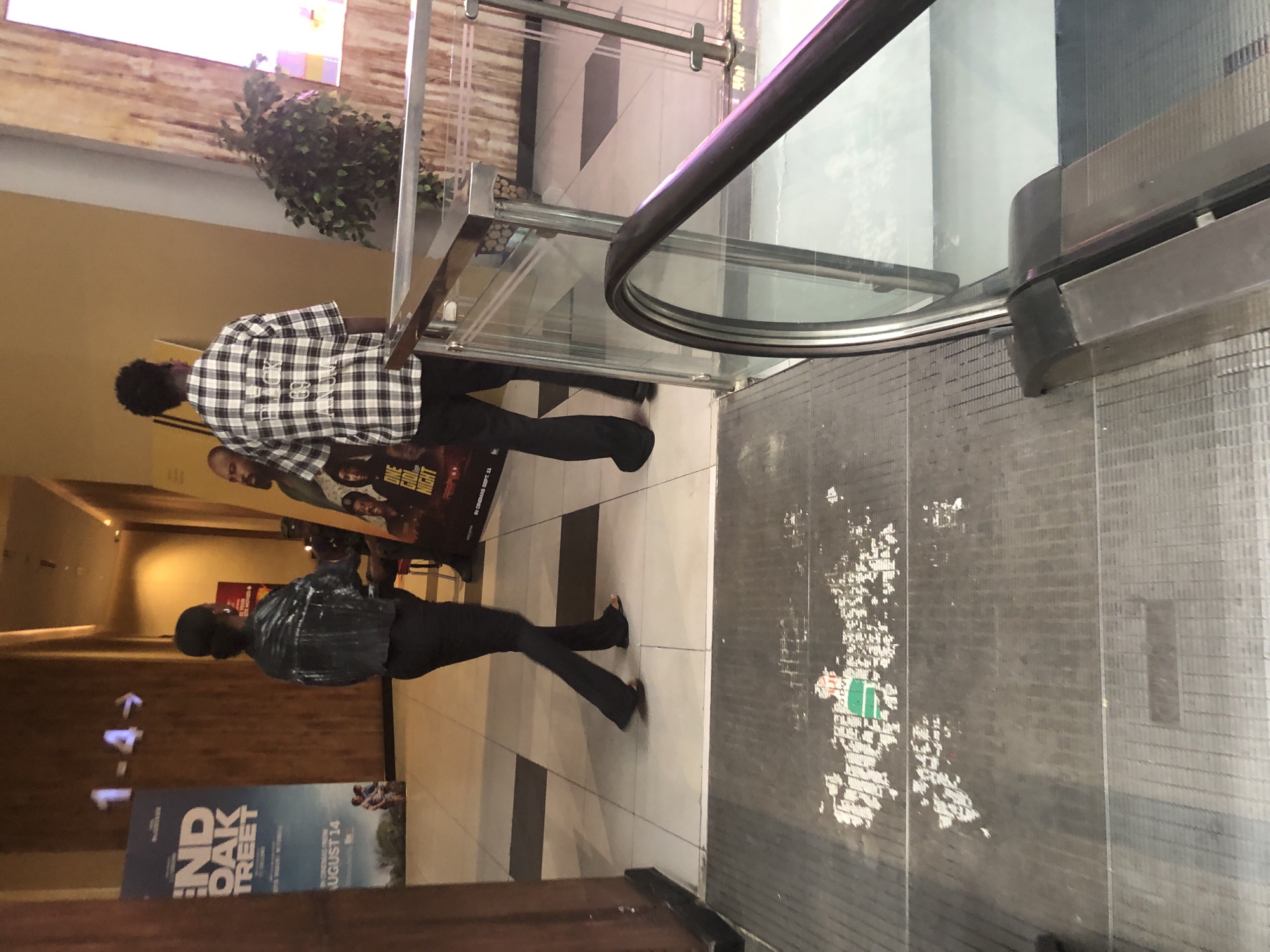The image depicts a hallway within a cinema complex. Two individuals are visible in the mid-ground, one facing away from the camera and wearing a black jacket and dark pants, and another facing away from the camera, wearing a black and white checkered shirt and dark pants. The hallway features movie posters on the wall, including one for "END OF OAK STREET" and another for "ONE GOOD NIGHT." Signage indicating seating rows "1-4" with arrows is present on the wall. A metallic escalator railing with glass panels is in the foreground, obscuring a portion of the scene. The floor appears to be tiled in a checkered pattern, with some areas showing wear and tear. The overall environment suggests a public space, likely a cinema lobby or hallway. The presence of specific movie posters and the indoor setting, combined with the architecture and flooring, are characteristic of a modern cinema in Sangotedo, Nigeria.