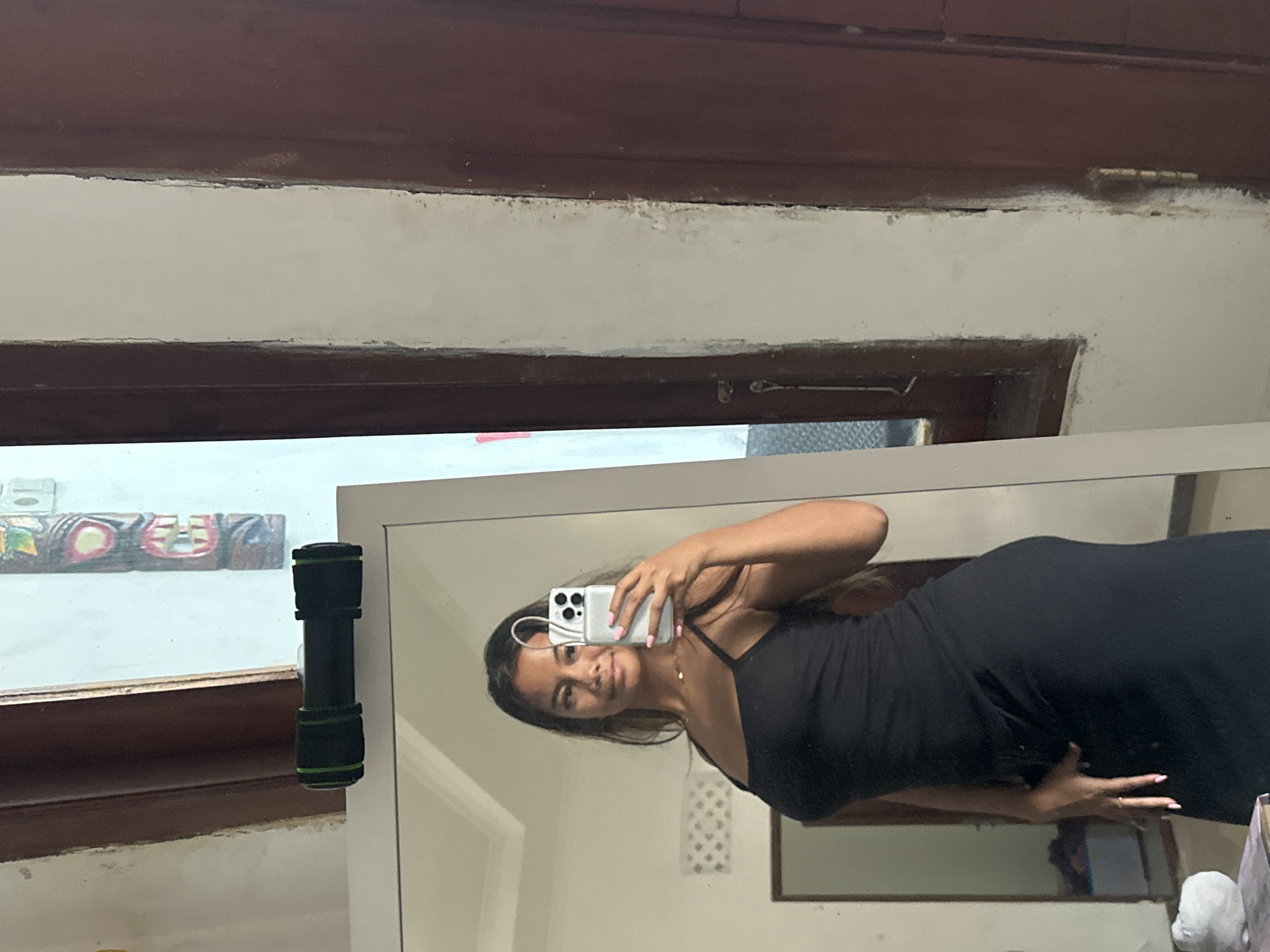 The photograph, taken inside what appears to be a dwelling in Sukawati, Indonesia, features a woman taking a selfie in a mirror. She is wearing a black dress. The room contains a window with a wooden frame and a flashlight is attached to the mirror. Outside the window, there are decorative wooden carvings. A partial view of the upper part of the wall and ceiling is also visible.