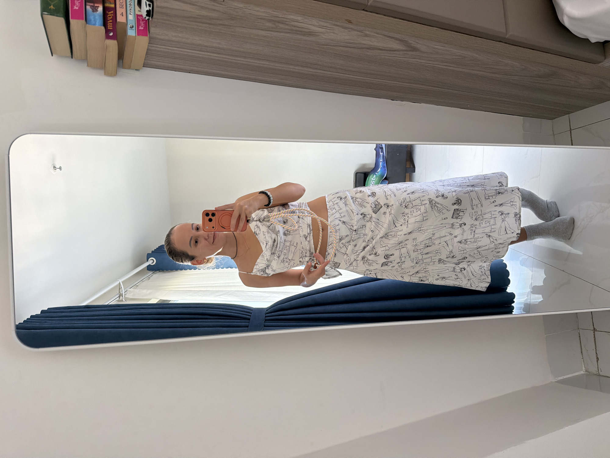 A young woman stands in front of a full-length mirror in a room. She is wearing a white two-piece outfit with a print of black line drawings. The top is a cowl-neck camisole with thin straps, and the skirt is a calf-length A-line skirt. She is holding a pink smartphone in her right hand, and a decorative rope is draped around her neck and shoulders. She is also wearing grey socks. The mirror reflects the woman, a dark blue curtain to her left, and a stack of books on a shelf to her right. The room appears to be a bedroom or a dressing area. The floor is tiled with white tiles, and the walls are painted white. The overall impression is of a personal space, likely a private residence in Nha Trang Ward, Vietnam.