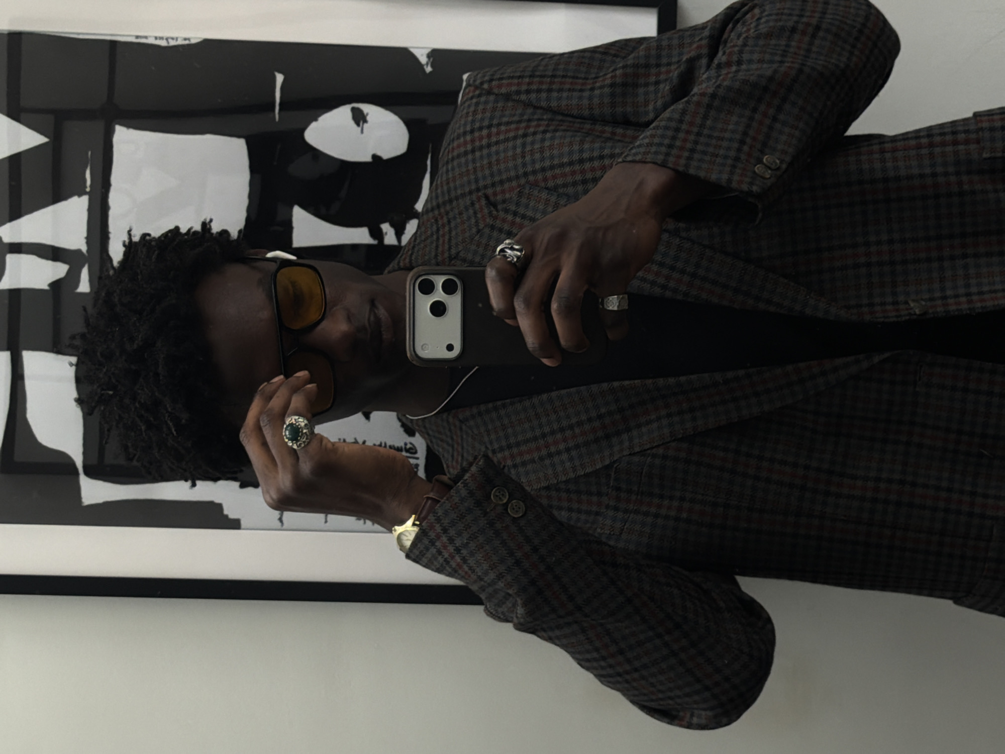 A person with dark skin is indoors taking a selfie with a smartphone. The person has dark hair styled in dreadlocks, is wearing brown sunglasses, and has several rings on their fingers. They are wearing a plaid blazer and a black shirt. A gold wristwatch is visible on their left wrist. Behind the person is a black and white abstract art print in a black frame, hanging on a white wall. The location is not discernible from the image.