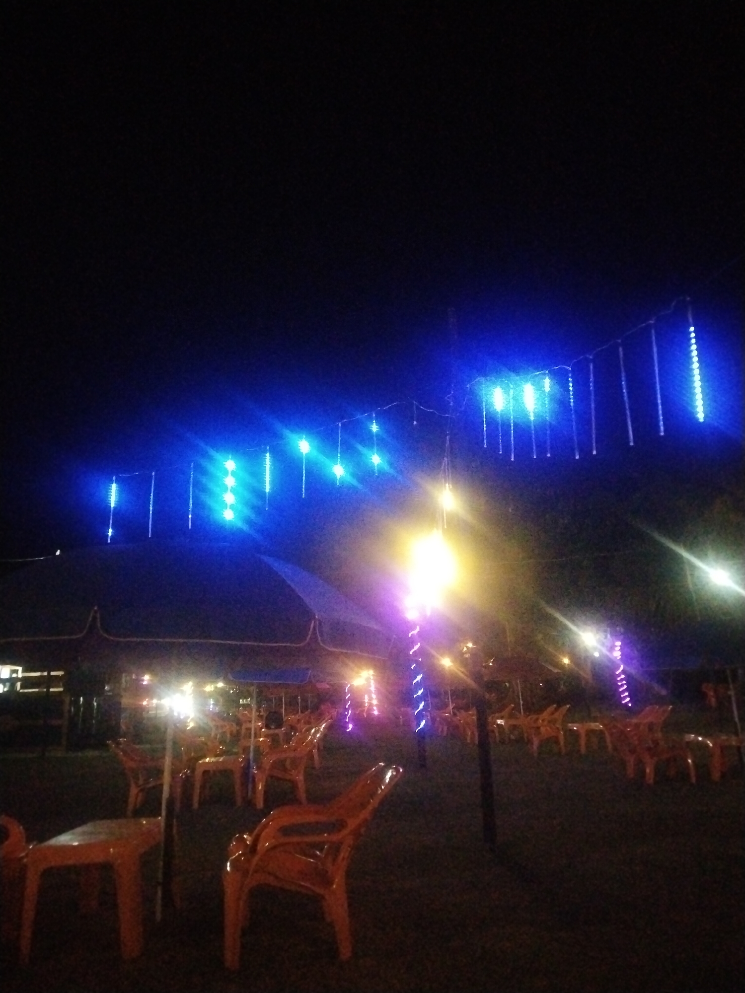 The image depicts an outdoor seating area at night in Glokaron, Indonesia. The scene is illuminated by a combination of hanging blue string lights and bright yellow-white light sources, one of which appears to be mounted on a pole wrapped with purple decorative lights. Orange plastic chairs and tables are arranged under what appears to be a large tent or canopy structure. The ground is dark, possibly grassy. No people are visible in the image.