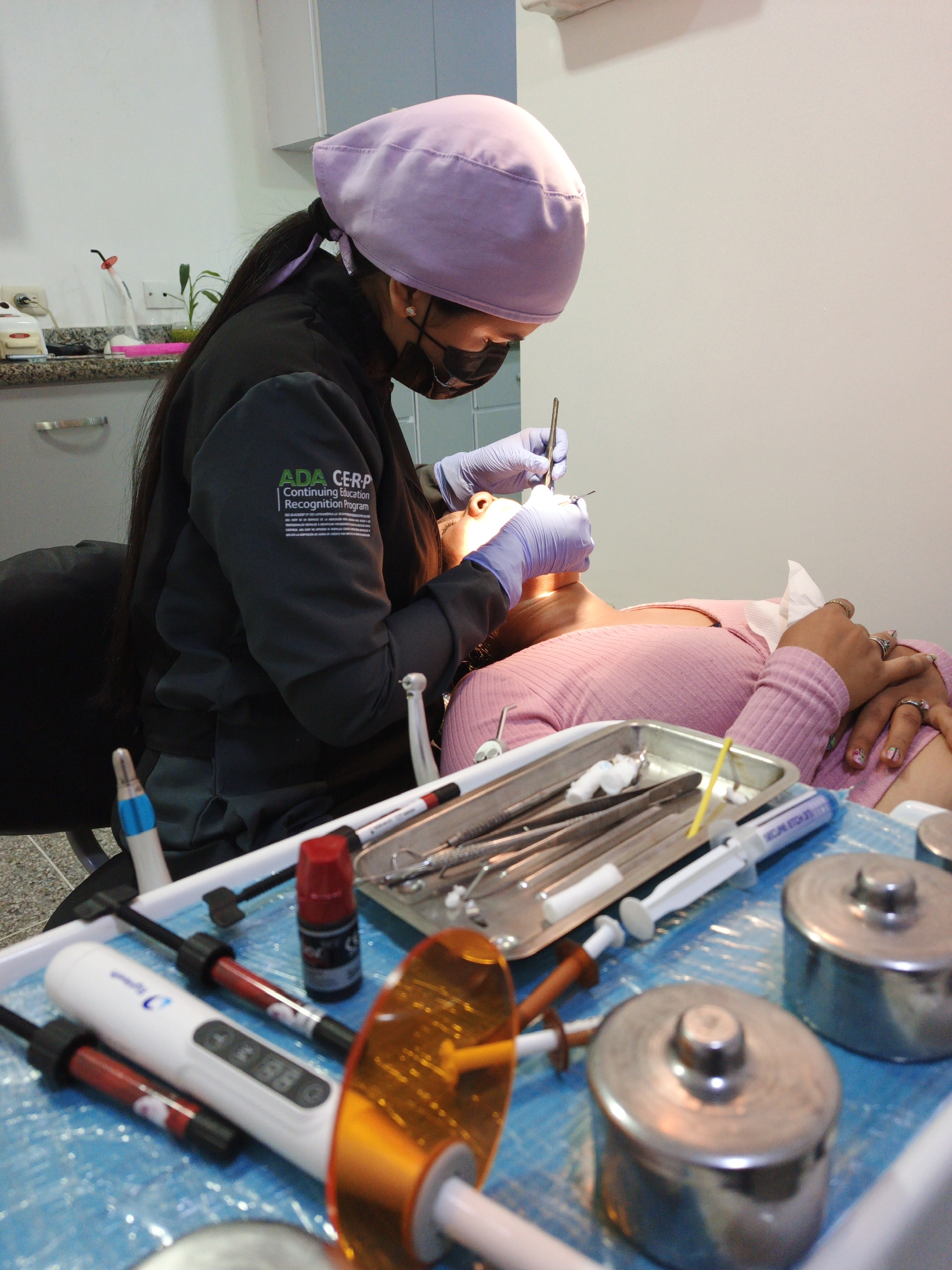 A dentist, wearing a lavender surgical cap, a black face mask, and blue latex gloves, is examining a patient. The dentist is wearing a black jacket with "ADA CERP Continuing Education Recognition Program" printed on the left arm. The patient is lying in a dental chair, wearing a pink sweater. On a tray table are various dental instruments, including tweezers, probes, syringes, and two small metal containers with lids. A red bottle and a white dental curing light with an amber filter tip are also on the tray. The location is a dental office in Tinaquillo, Venezuela.