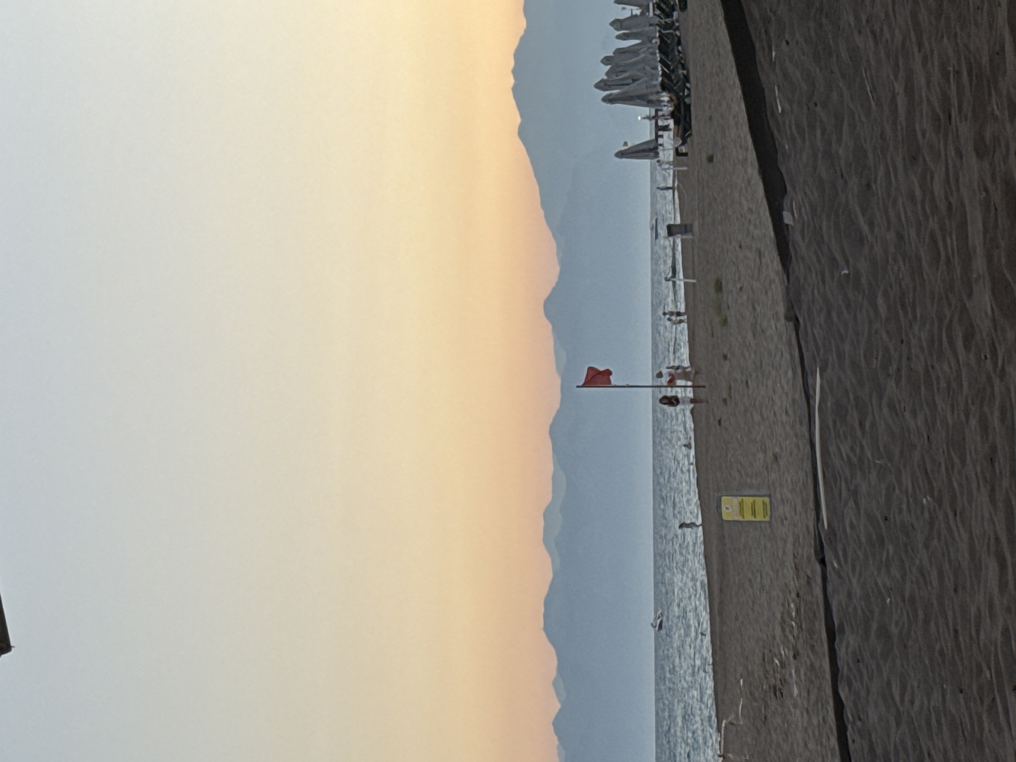 The image depicts a beach scene at what appears to be sunrise or sunset, given the soft, warm tones of the sky. The sky transitions from a pale yellow-orange near the horizon to a light blue. In the distance, the silhouettes of mountains are visible, creating a layered effect against the sky.
The beach itself is dark, with fine, textured sand. Along the edge of the beach, closer to the water, are several rows of closed beach umbrellas and lounge chairs, suggesting a resort or public beach area. A red flag is planted on a pole near the water's edge, possibly indicating a warning or a change in swimming conditions.
A few individuals are present on the beach. Some appear to be standing near the water, while others are further back, possibly near the umbrella setup. Their presence is subtle, integrated into the broader scene. The sea is calm, with gentle ripples reflecting the light.
A yellow sign with some text is affixed to a pole on the beach, but the text is not legible. The overall mood is tranquil and serene, characteristic of a quiet moment by the sea. The location is identified as Serik, Turkey.