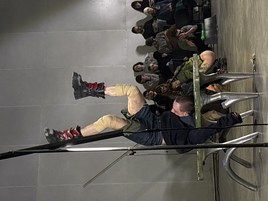 This image captures an indoor performance event occurring in Sofia, Bulgaria. The central focus is on two individuals interacting with a low, green-painted wooden platform featuring stylized silver-colored, curved legs.
One performer is positioned inverted, with their legs extended upwards. They wear dark shorts, beige wraps around their knees and shins, and heavy black boots with red accents on the heels/soles. Their upper body is below the platform, with their head, featuring short hair and a beard, resting horizontally on the platform surface, facing left. A black vertical pole, possibly a light stand, obstructs part of their left leg on the left side of the frame.
A second performer is partially visible underneath the platform, appearing to be in a similar or complementary inverted position, with beige-wrapped legs and light-colored hair discernible.
In the background, approximately twenty-five individuals are seated in multiple rows on dark folding chairs, observing the performance. The background consists of a light grey, paneled wall and a light-colored floor, under uniform illumination.