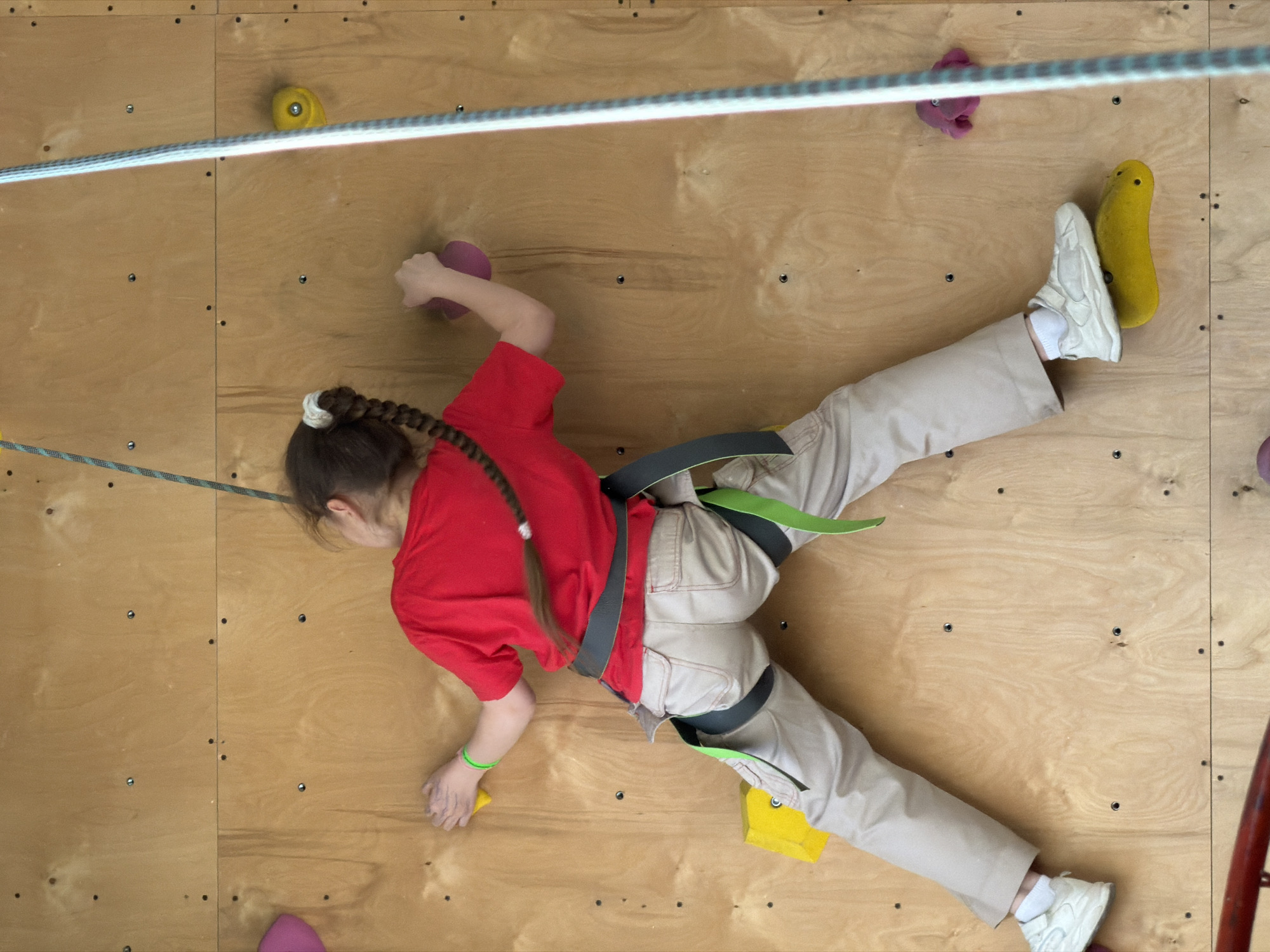 A girl is climbing an indoor rock climbing wall, harnessed for safety. She wears a red t-shirt, beige pants, and white sneakers. Her hair is pulled back in a braid. She is using various colored hand and foot holds on the plywood wall. A safety rope runs across the top of the wall. The location is Izhevsk, Russia.