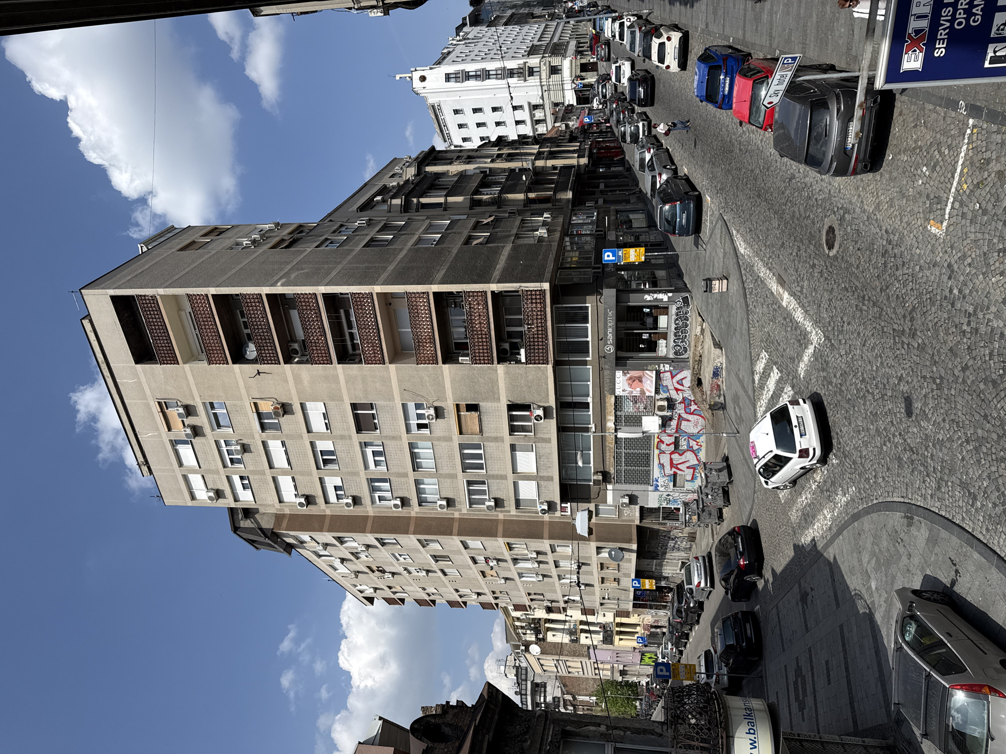 This daytime photograph captures a vibrant urban street scene in Belgrade, Serbia, on a clear, sunny day with a bright blue sky dotted with white clouds. The street, partially paved with cobblestones and partially with asphalt, curves gently from the foreground towards the distance, lined with numerous parked cars of various models and colors including red, blue, grey, white, and black. A white car is visible turning in the middle ground, while a dark grey SUV is parked prominently near a street sign.
On the left side of the street stands a tall, multi-story concrete apartment building, characteristic of Brutalist or post-war Eastern European architecture. It features numerous windows, balconies with patterned railings, and many visible air conditioning units attached to its exterior walls. Further down the street on the right, a distinctive white building with numerous windows and a more classical facade can be seen. Smaller commercial buildings and residential structures line both sides of the thoroughfare, some featuring graffiti.
A few pedestrians are visible on the sidewalks, adding to the sense of everyday activity. Visible text provides location cues: a prominent "Sky hotel P" sign, indicating a hotel and parking, and various other signs like "SanioPTIK" and "w.balkans". A Serbian license plate, "BG 2553 SH", clearly identifies the city as Belgrade ("BG"). Another sign partially reads "EXTR SERVIS OPR GAM," likely advertising extra services, repairs, or games. The scene conveys a bustling city environment under bright sunlight, with shadows indicating mid-day.