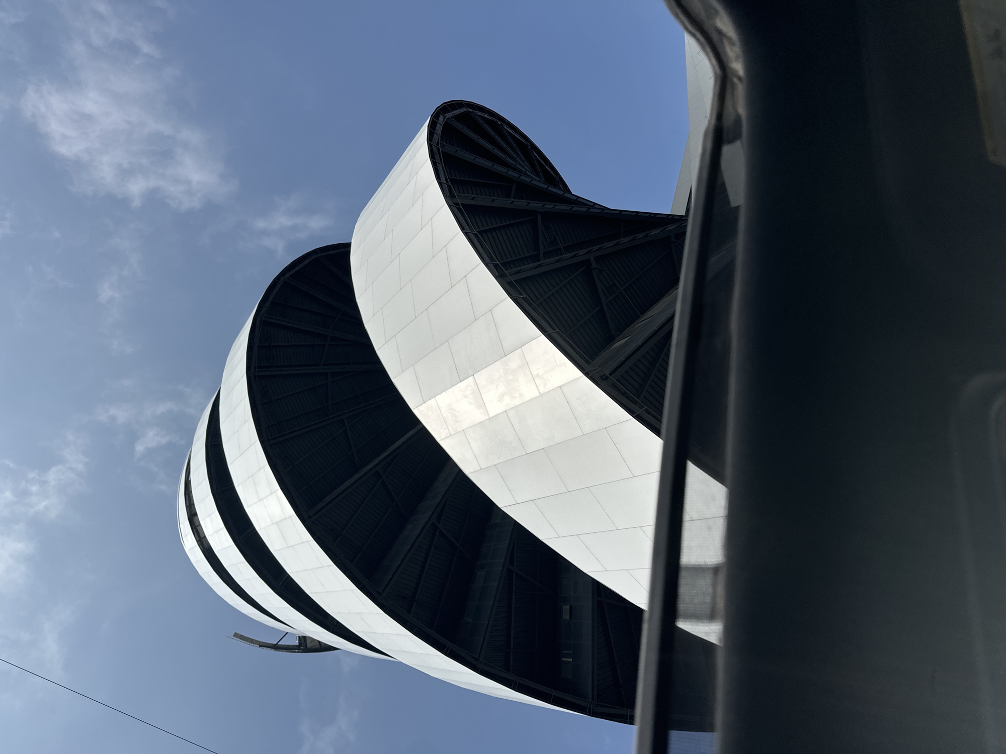 The image shows a portion of the exterior architecture of the Museo Internacional del Barroco (International Baroque Museum) located in San Bernardino Tlaxcalancingo, Mexico. The photograph is taken from a low angle, looking up at the museum's distinctive spiraling facade. The facade is comprised of alternating black and white horizontal bands. The sky is visible in the background, with some wispy clouds. A gray vertical structure, potentially part of a vehicle, is visible on the right side of the image, partially obstructing the view of the museum. There are no people visible in the frame.