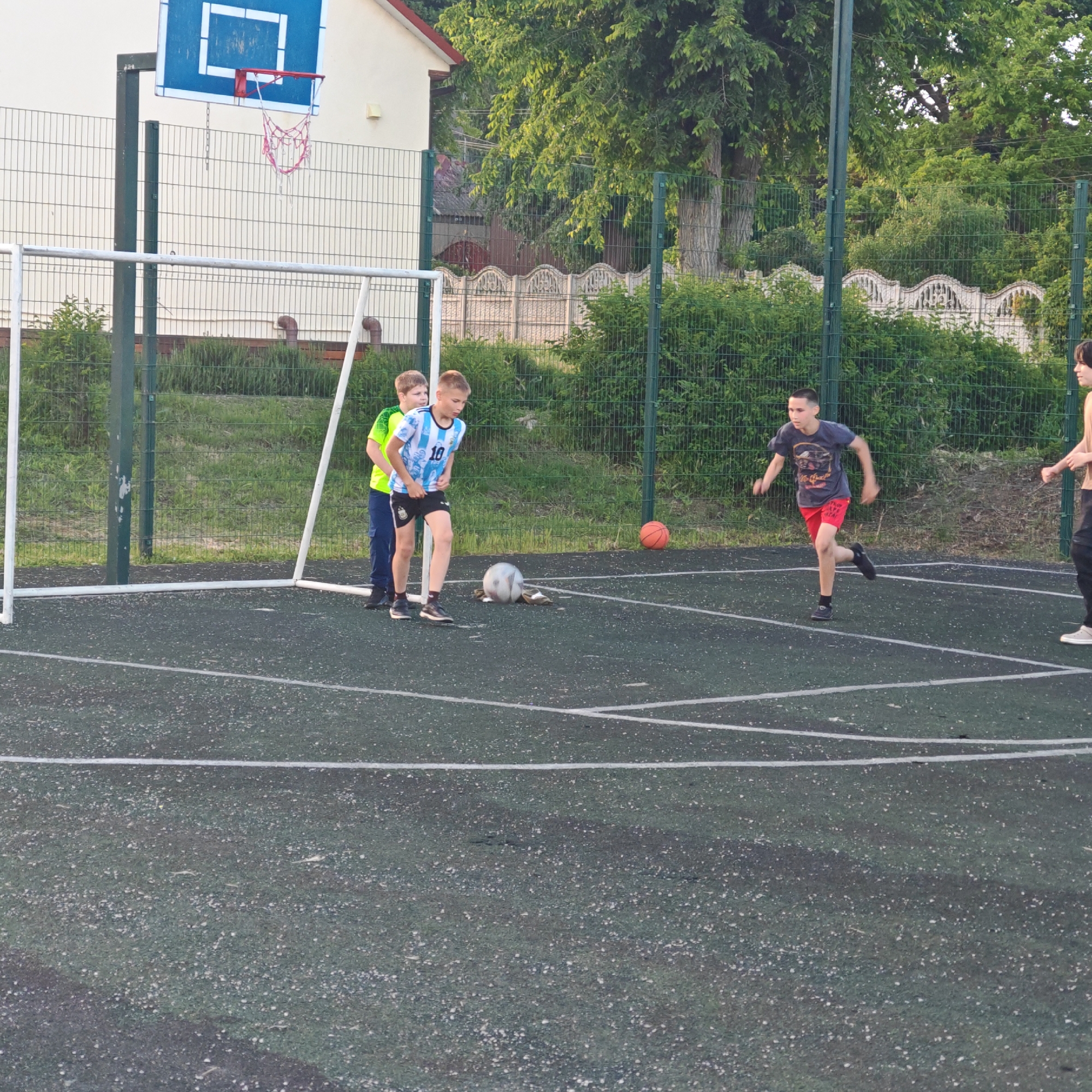 The image depicts a group of boys playing what appears to be a game of soccer in an outdoor recreational area located in Мысовское сельское поселение, Ukraine. The setting includes a combination soccer and basketball court. A soccer goal is visible on one end of the court, and a basketball hoop is situated in the background. Several children are in the foreground of the image engaged in the activity. One boy in a blue and white Argentina jersey appears ready to kick a soccer ball. Another boy is running in the opposite direction. The surrounding environment is characterized by a green fence, trees, and buildings, suggesting a park or recreational facility. The time of day seems to be in the late afternoon or early evening.
