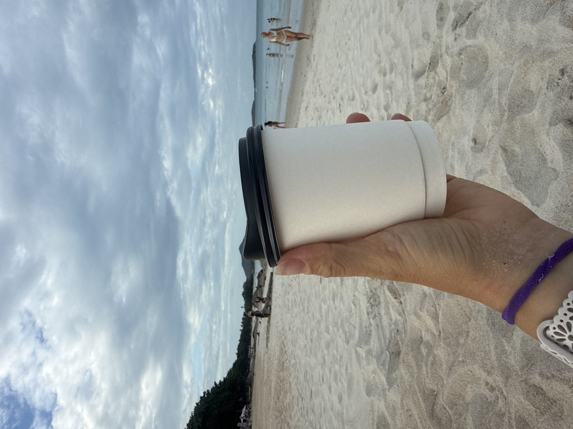 A close-up, first-person perspective shot shows a hand holding a white paper coffee cup with a black lid on a sandy beach. The cup is the primary subject, with its texture and plain white surface clearly visible. In the background, the beach stretches out, with the ocean visible and several people in the distance. Some individuals are wading in the water, while others are walking along the shoreline. The sky is overcast with clouds. The scene suggests a relaxed atmosphere on a beach, potentially in a tropical location like Ko Samui District, Thailand, given the typical beach settings and vegetation seen.