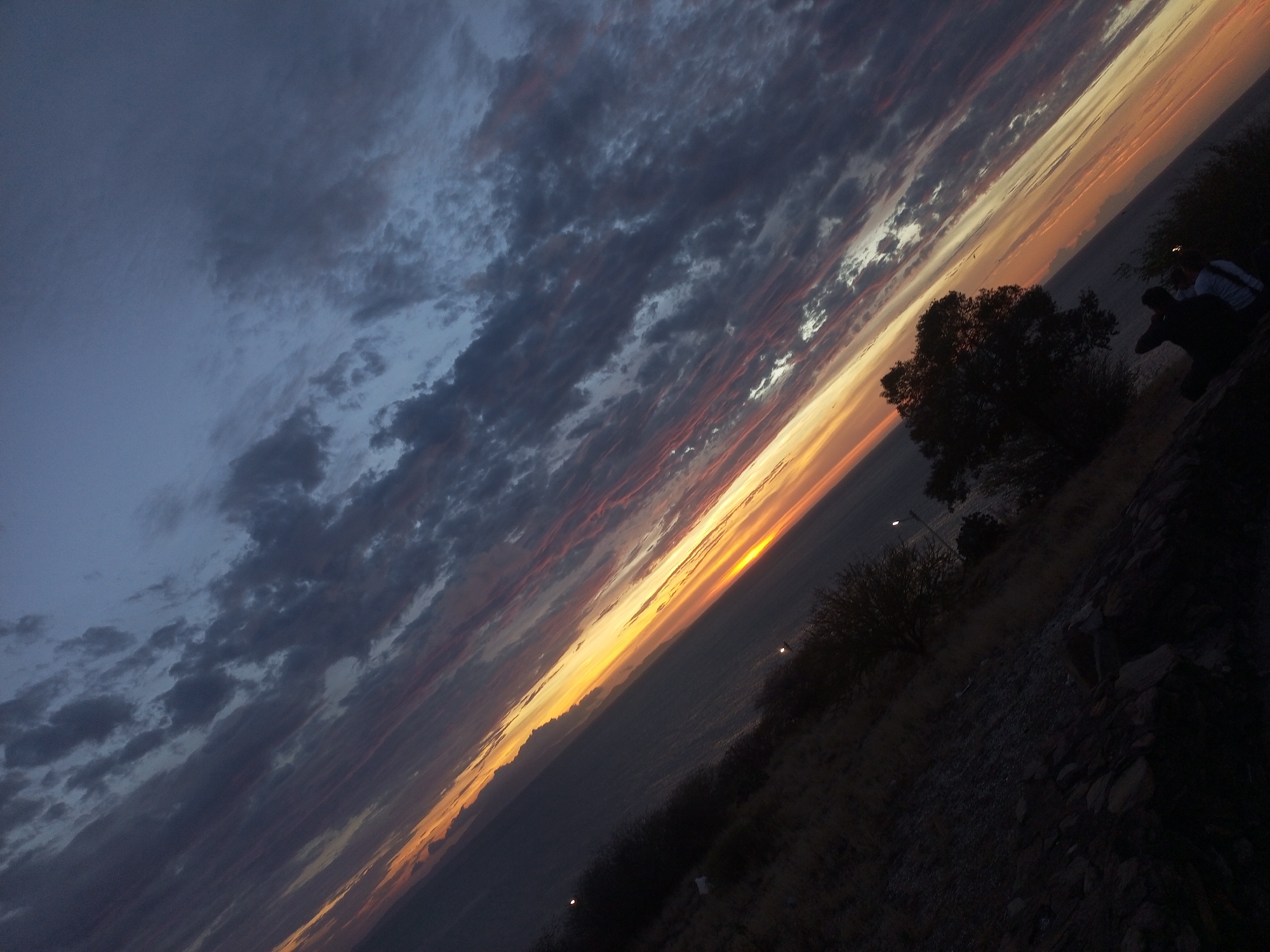 The image shows a twilight or sunset scene overlooking a city landscape. The sky, a dominant feature, transitions from a dark blue-gray with scattered clouds on the left to a vibrant orange and yellow band on the horizon, indicating the sunset. The city lies beneath the orange band, appearing as a dark, indistinct area. In the foreground, a hillside is visible, covered with sparse vegetation and rocks. On the right side, a group of people are silhouetted against the sky, appearing to be observing the sunset. The angle suggests that the image was taken at an incline. Based on the location of Juan Griego, Venezuela, the sunset is likely seen over the coast, with the city sprawling along the coastal landscape.