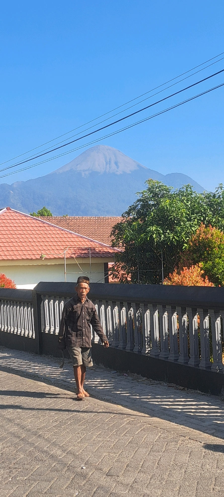 A full-body shot captures an elderly man walking on a paved sidewalk in what appears to be a residential area in Sekarjoho, Indonesia. The man is wearing a dark, long-sleeved plaid shirt, grey shorts, and a dark, patterned cap. He is walking towards the left of the frame, with his left leg extended forward. He has a dark object, possibly a bag, hanging from his left shoulder. The sidewalk is made of grey interlocking bricks and is partially shaded by a decorative wall with white balusters. Behind the wall, there are houses with red tiled roofs and green trees. In the background, a large, pale mountain looms under a clear blue sky. Overhead, power lines stretch across the sky. The lighting suggests it is daytime, with strong sunlight casting distinct shadows.