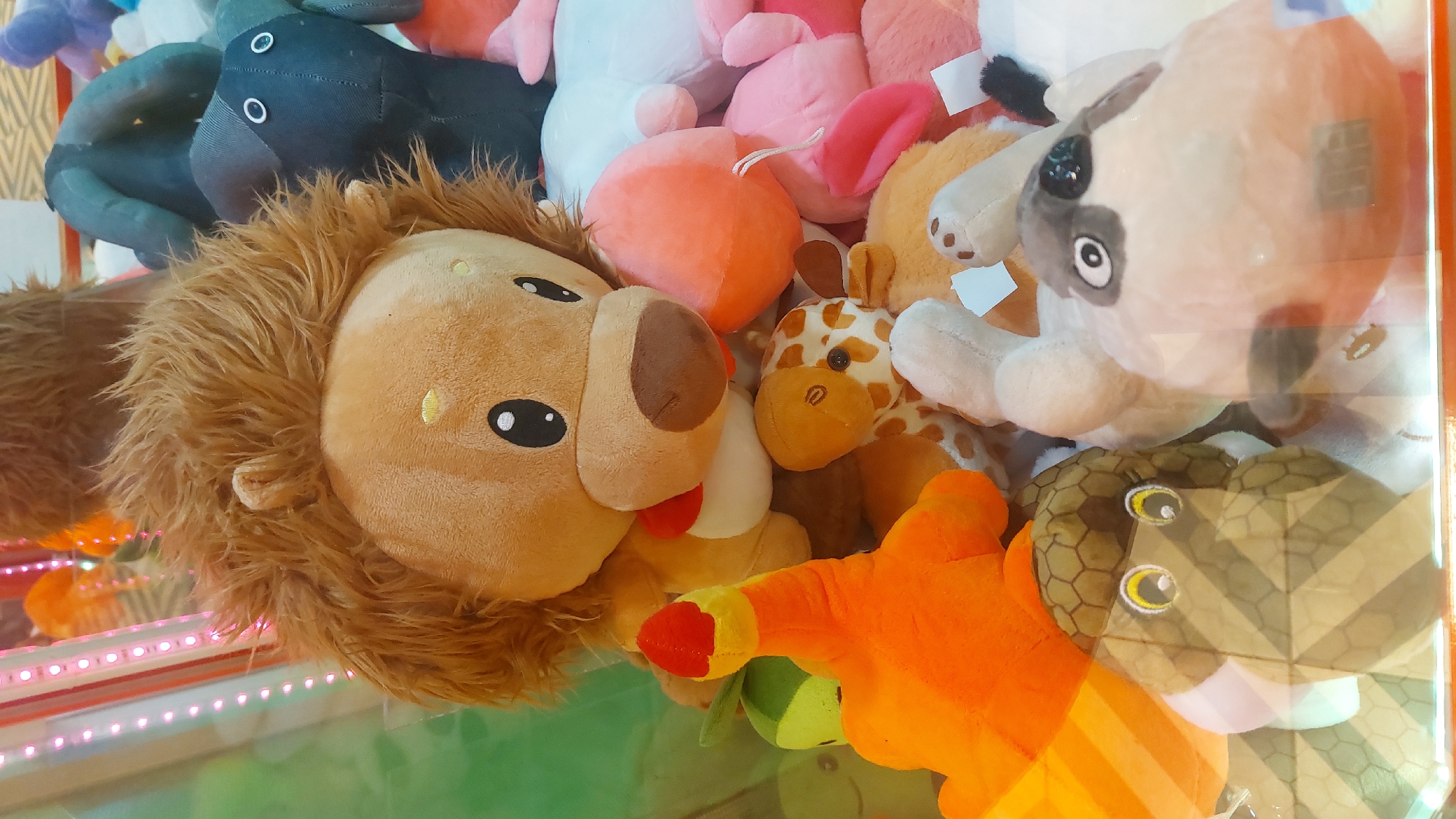The image depicts a claw machine filled with plush toys. The machine contains a lion plush with a brown mane, a giraffe plush, a snake plush with a hexagonal pattern, a dog plush with black markings, an orange claw with a red tip, and other indiscernible plush toys. The machine appears to be located in Novosibirsk, Russia, as it was retrieved from there. The image provides no information about people, actions, or interactions. The environment appears to be an indoor arcade or entertainment venue.