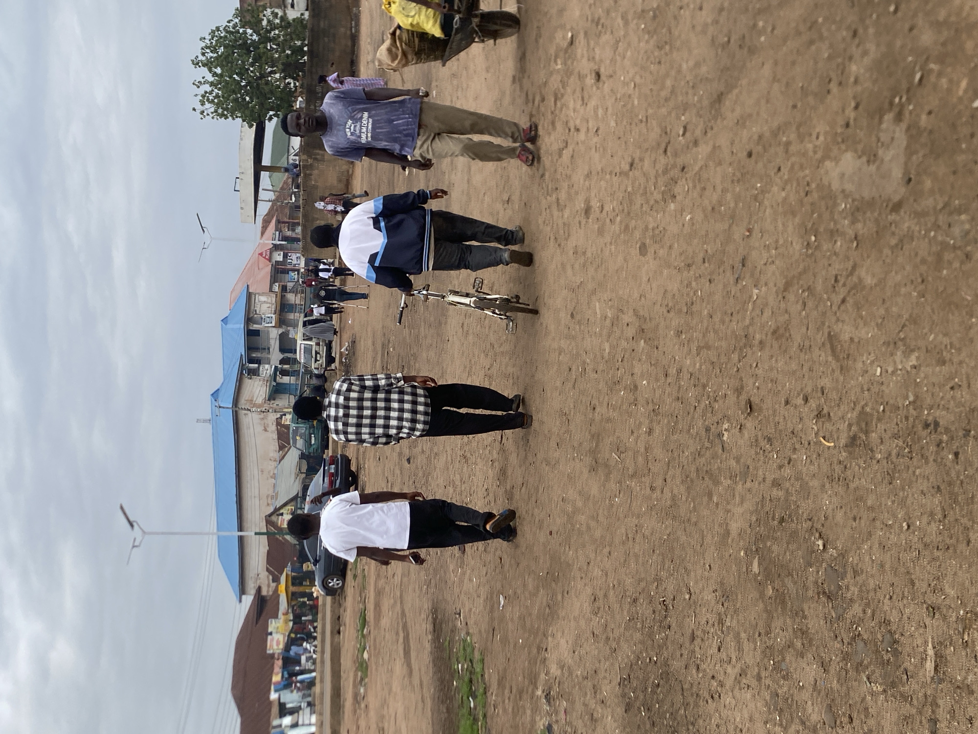 The image depicts an unpaved road in Bukuru, Nigeria. Four individuals are walking in the same direction. The first man is wearing a purple shirt and khaki pants. A second man is walking alongside a bicycle. The third man wears a black and white checkered shirt and black pants. The fourth man is in a white shirt and black pants. Buildings and trees are visible in the background. A lamp post is also visible. The sky is overcast.