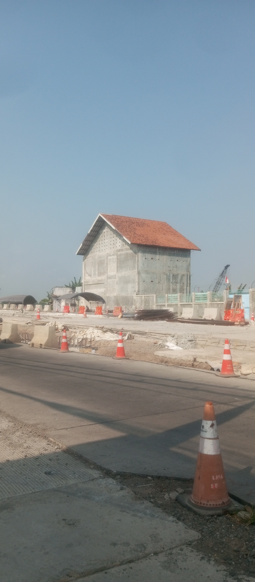The image depicts an outdoor scene under a clear blue sky, likely in Kediri City, Indonesia, given the context. In the mid-ground, a large, unfinished concrete building with a terracotta tile roof stands prominently. The building has a high-pitched roof and several rows of small, circular openings on its upper facade. To the left of the building, several structures with arched, covered roofs are visible, possibly part of a construction site or temporary shelters.
The foreground and mid-ground are dominated by a road under construction. Orange traffic cones, some with white reflective bands, are strategically placed to delineate work areas and guide traffic. Several concrete barriers are also visible, indicating ongoing roadwork. Piles of construction materials, including rebar and what appears to be gravel, are scattered throughout the site. A large construction crane, with a red and white flag attached, is visible in the background to the right of the concrete building. The ground surface consists of asphalt and concrete, with some areas showing exposed soil and gravel. The overall impression is one of an active construction zone.