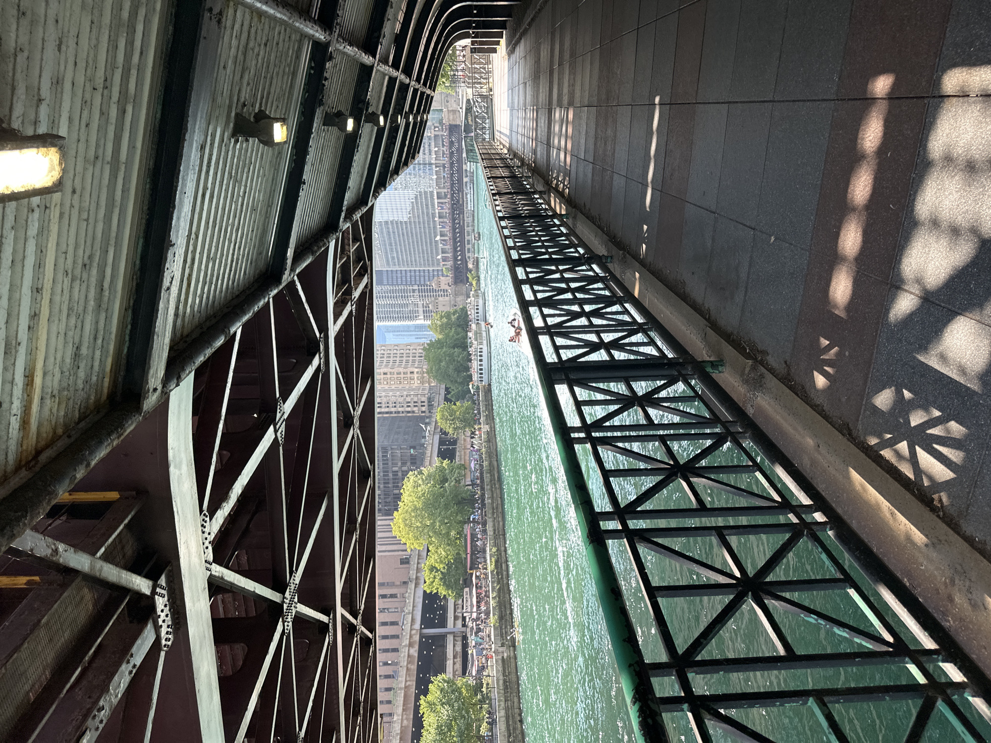 The image depicts a scenic view of the Chicago Riverwalk on a sunny day. The perspective is from under a covered walkway, looking out towards the river and the city skyline. The walkway itself is paved with large, square tiles, interspersed with reddish-brown rectangular tiles, and features a metal railing along the edge overlooking the river. The railing is black with green accents at the posts and has a geometric pattern. Sunlight casts intricate shadows from the railing onto the walkway.
The river is a vibrant emerald green, with several boats visible, including tour boats and smaller personal craft. People can be seen on the riverbanks, enjoying the day. Lush green trees line the opposite bank, providing a natural contrast to the urban landscape of tall buildings in the background. The architecture of the city is visible, including bridges spanning the river and the distinctive facades of skyscrapers. Above, the underside of a bridge structure is apparent, composed of a complex network of metal beams and supports. The walkway also has a corrugated metal ceiling with industrial-style lighting fixtures. The overall scene conveys a lively, urban environment with a strong connection to its waterfront.