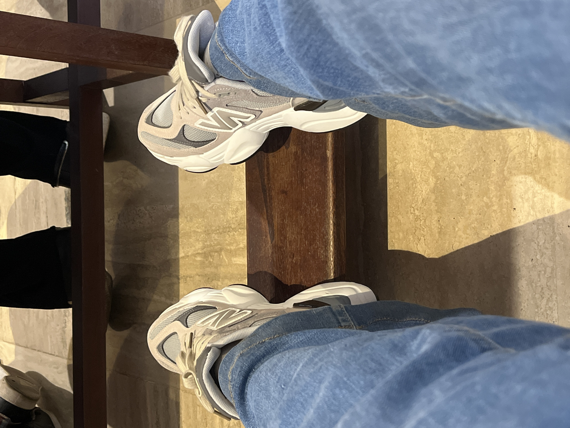 The scene, located in Santa Ana, Venezuela, is depicted from a first-person, downward-looking perspective, focusing on an individual's lower extremities. The subject wears light blue denim jeans and a pair of New Balance athletic shoes. These shoes are characterized by prominent white, sculpted soles, light beige and gray mesh uppers, and the visible 'N' logo on the side of the right shoe. The shoes are resting with their soles flat against a horizontal, dark brown wooden footrest, which appears to be part of a larger wooden furniture structure; a vertical wooden support is visible on the right side of the frame. The floor beneath and extending into the background is composed of light-toned, textured tiles, possibly marble or ceramic. In the upper background, partially obscured by the wooden footrest, the lower legs and feet of two additional individuals are visible: one on the left wearing dark trousers and dark shoes with a white sole, and another on the right in dark trousers and dark shoes. The lighting suggests an indoor environment, casting distinct shadows from the feet and furniture onto the tiled floor.
