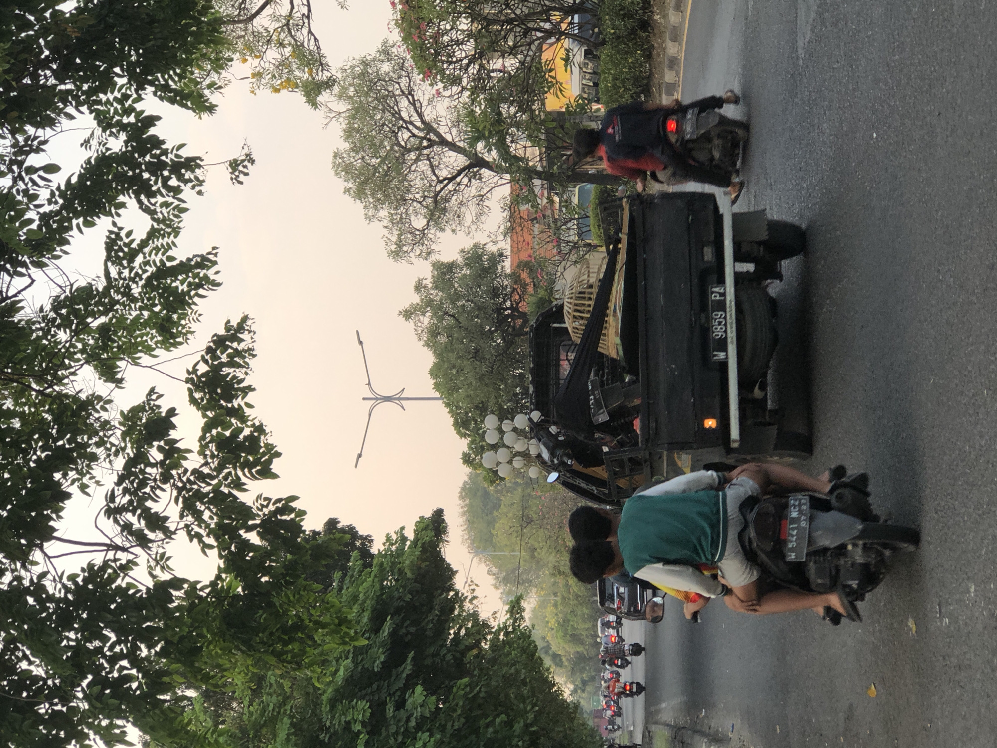 A street scene in Sumorame, Indonesia, captures a moderately trafficked road lined with trees. In the foreground, two young boys, one wearing a green and white jacket and the other a yellow shirt, ride on the back of a motorcycle. Their legs are visible, and they appear to be looking forward. The motorcycle is in motion, as indicated by the blur of the wheels and the general context of traffic.
In front of the motorcycle, a black pickup truck is visible. The truck is carrying several items in its bed, including what appears to be a birdcage and some wooden planks. The truck's license plate reads "W 9859 PA." To the right of the truck, another motorcycle carrying a single rider wearing a red and black shirt is also in motion. The rider's legs and feet, clad in flip-flops, are visible.
The road surface is asphalt, with some visible wear and tear. On the right side of the road, there is a sidewalk and vegetation, including flowering trees with pink and yellow blossoms. In the background, there are more trees, buildings with red roofs, and a street light with an ornate design. The sky is a hazy, light orange, suggesting either sunrise or sunset, and a light pole with a decorative structure is visible in the distance. The overall impression is a snapshot of daily life in a bustling Indonesian city.
