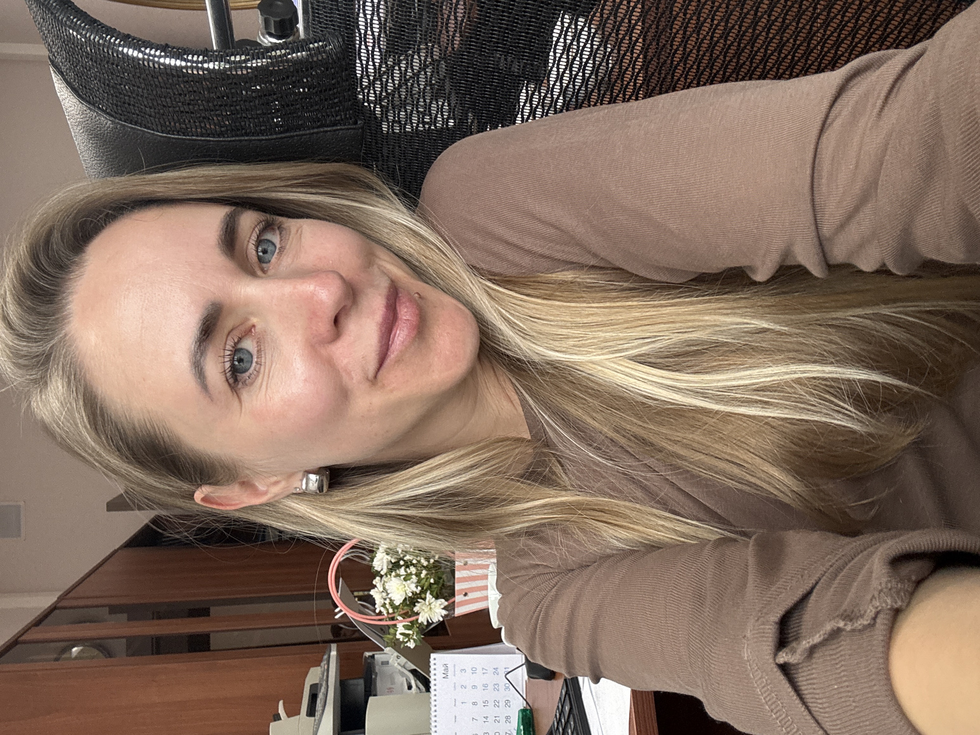 A woman with blonde hair and blue eyes is positioned in a black mesh office chair. She is wearing a brown long-sleeved shirt. Her face is close to the camera. Behind her, there is an office desk with a calendar displaying the month of May in Russian. White flowers are also visible on the desk. A wooden cabinet is partially visible in the background. The photo was taken in the city of Chaikovsky, Russia.