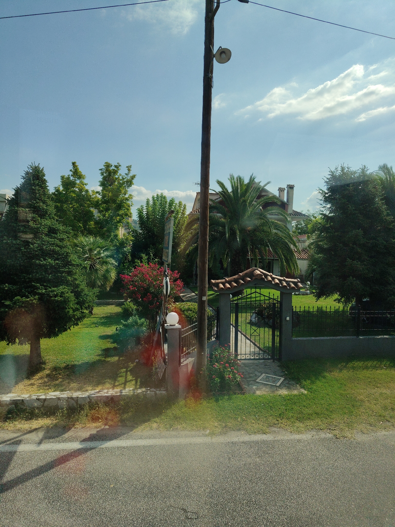 The image shows a residential property entrance with a gate in Vasiliki, Greece. The gate is made of black metal with a curved, tiled roof above it. It opens onto a well-maintained green lawn. On the left side of the property, there are several trees, including a large evergreen tree and a palm tree. A wooden utility pole with a light fixture is positioned to the left of the gate. In the background, parts of a house with a tiled roof and chimneys are visible. The sky is clear with some scattered clouds. The foreground shows a paved road with a white line. The overall scene depicts a sunny day in a suburban or rural setting.