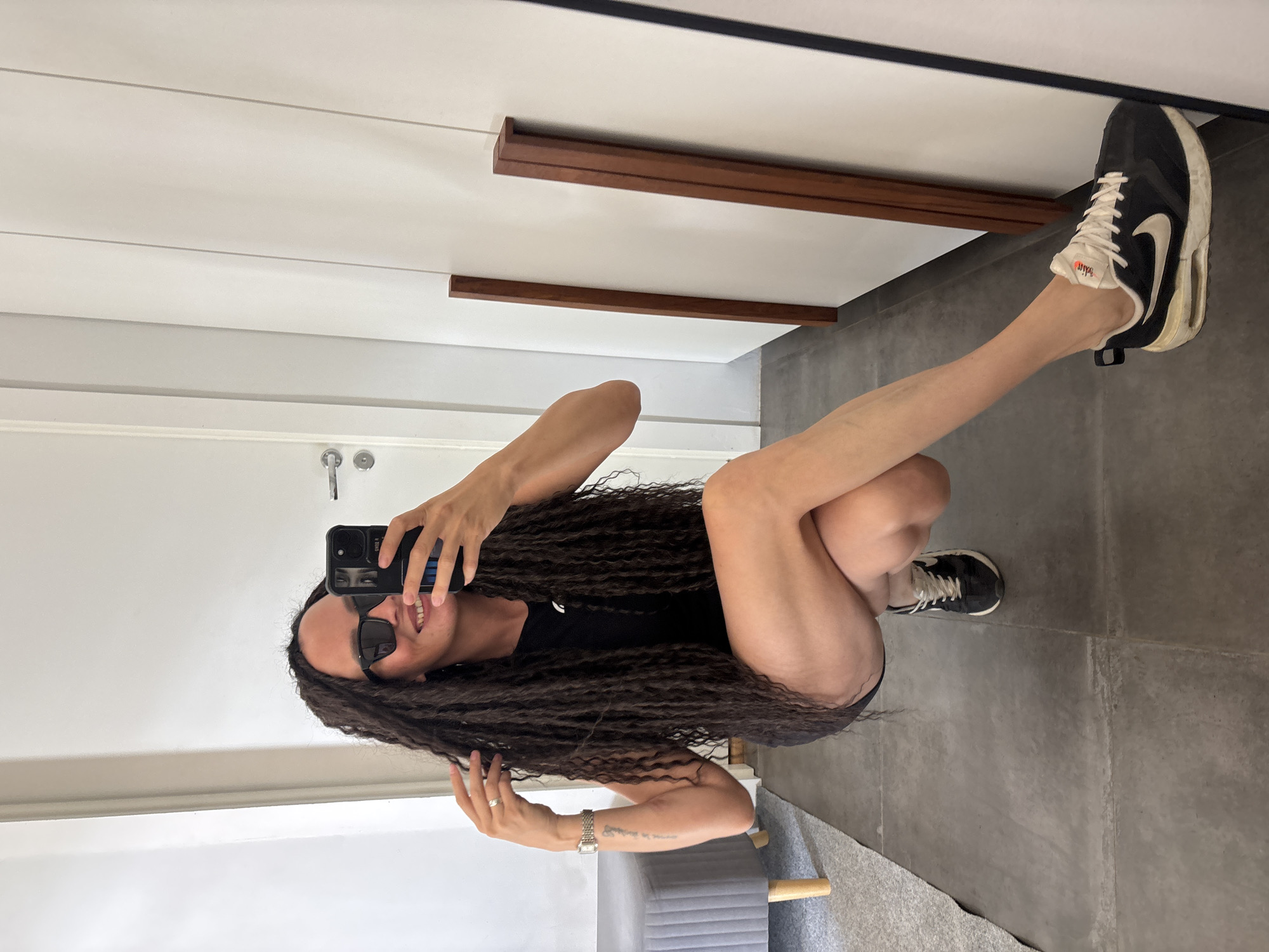 A woman is crouched down, taking a picture of herself in a mirror. She is wearing sunglasses and a black outfit. She has long, dark, curly hair. She is holding a black smartphone in her left hand. Her right hand is raised near her hair. She is wearing a watch and a ring. She is wearing black and white sneakers. The room appears to be a dressing room or closet with a white wardrobe and gray tiled floor. A gray ottoman is behind her. There is a door with a silver handle in the background. It is not possible to confirm the location as Chaikovsky, Russia from the visual information.