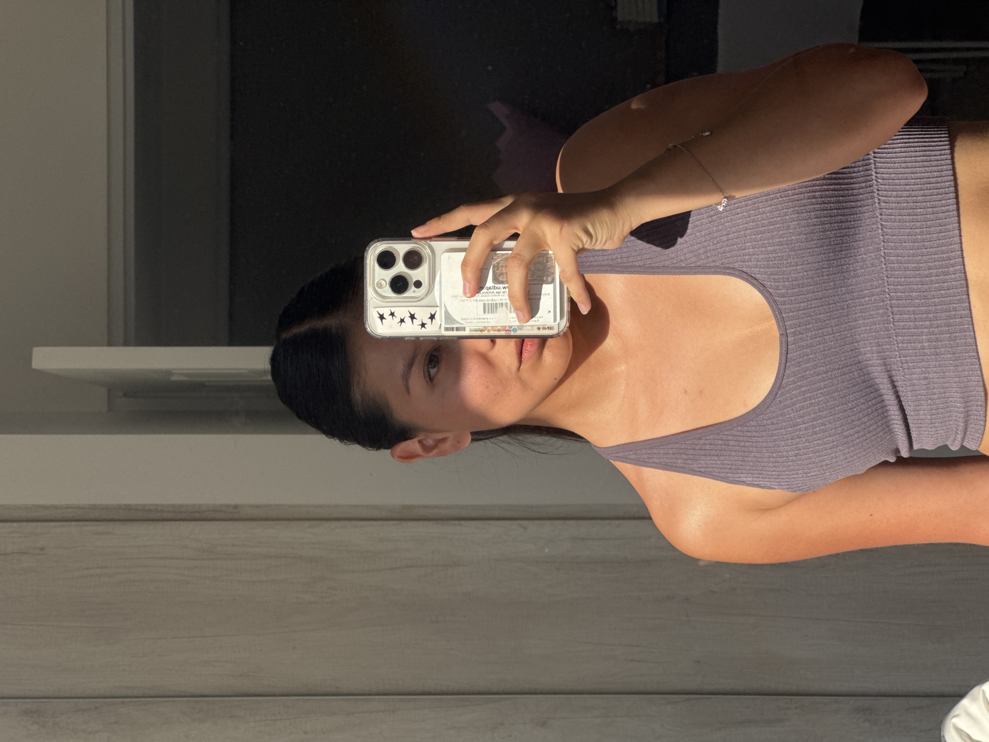 The image features a person, likely female, taking a selfie while holding an iPhone in a clear case that partially obscures their face. They have dark hair and are wearing a ribbed, muted lilac or purple tank top. A delicate, silver-toned bracelet with small charms is visible on their wrist.
The smartphone, identified by its triple-lens camera system, has several items tucked into its clear case. These include what appear to be two cards, one with a barcode and another with legible, mirrored text that seems to read "qiupibu.w" along with other indiscernible characters. A strip of black star stickers is also visible behind the phone.
Strong, directional sunlight illuminates the person's left arm and parts of their face, creating sharp shadows that cut across their features. The background is divided between a dark, shadowy area (possibly a window or dark wall) and a light-colored horizontal surface or shelf. Below this, a textured surface resembling light wood paneling is visible. The overall lighting suggests either early morning or late afternoon. The person's expression is neutral, with only one eye and parts of their mouth visible due to the phone's placement. The image is rotated, presenting the subject horizontally within a portrait orientation.