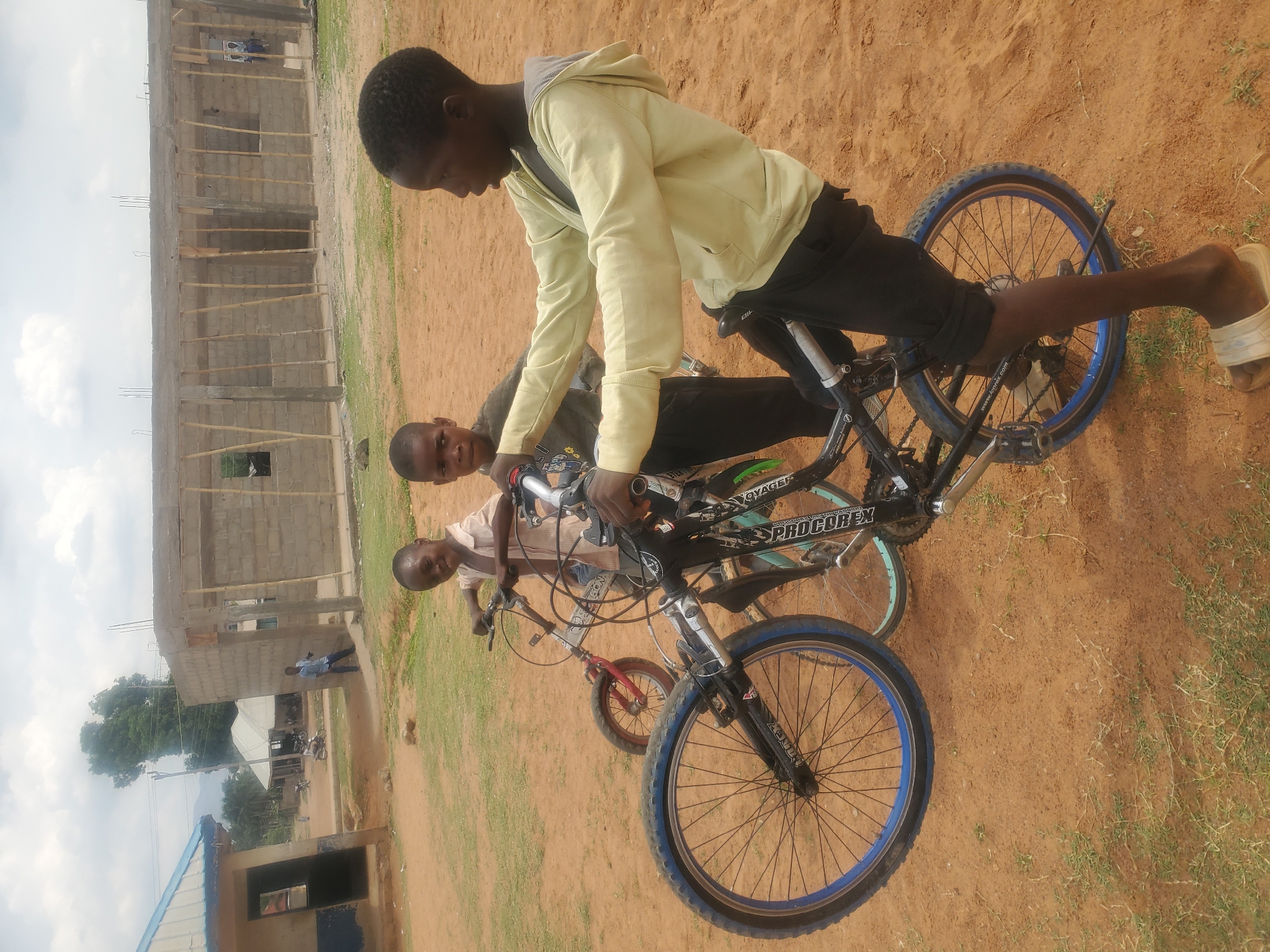 The media file captures an outdoor scene in Akwanga, Nigeria, featuring three young males with bicycles against a backdrop of buildings and open ground.
The central figure, a young male wearing a light yellow hooded sweatshirt and dark trousers, is mounted on a black bicycle with blue-rimmed wheels. His right foot is placed on the ground, wearing a white sandal. He is looking down and to his left. Behind him, another young male in a grey shirt is also on a bicycle, looking towards the camera. To the right and slightly in front of them, a third, younger boy wearing a light-colored striped shirt holds onto a smaller, red bicycle, looking directly at the viewer with a slight smile.
The ground is predominantly reddish-brown dirt, with scattered patches of green grass, suggesting a dry or semi-arid environment. In the background, a large, unfinished building constructed from concrete blocks is visible, with wooden scaffolding or support beams in place. Further in the distance, another building with a blue roof and some trees can be seen under a cloudy, overcast sky.
Visible text on the side of the main bicycle frame includes "PROCOREX" and "VOYAGER". There might also be "www.longtex.com" visible on a tire or rim, but it is faint. The overall scene depicts a casual, possibly recreational moment among the boys in what appears to be a developing or semi-urban area.