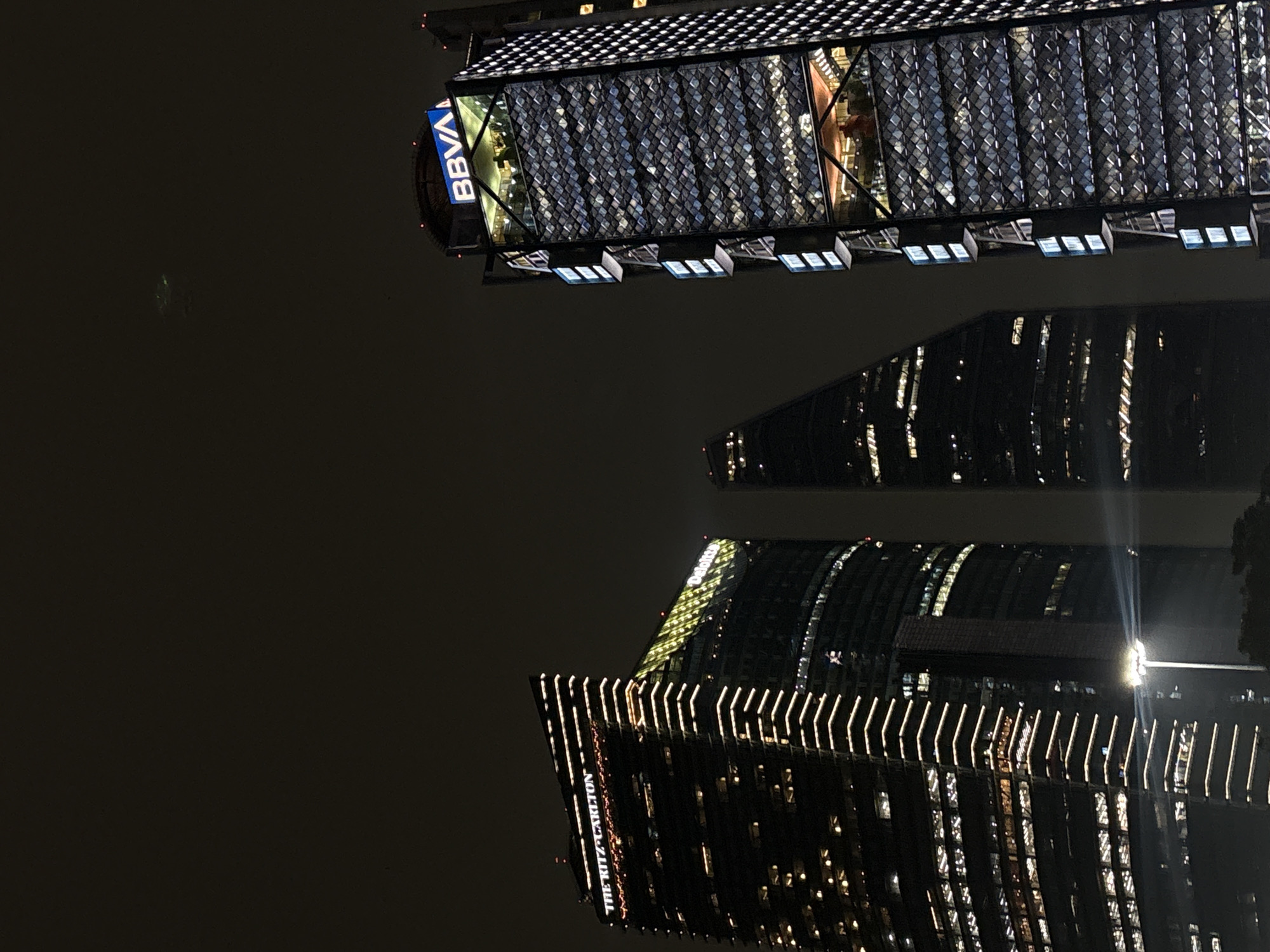 This nocturnal photograph captures a street-level view of three prominent skyscrapers along Paseo de la Reforma in Mexico City, Mexico. The building on the left is the Chapultepec Uno R509 tower, displaying illuminated "THE RITZ-CARLTON" signage near its top and "Deloitte" signage below, with distinctive horizontal light strips outlining its structure and various illuminated windows. To its immediate right, partially obscured, stands a darker, triangular-shaped skyscraper, identified as Torre Reforma, with scattered illuminated windows. On the far right, the Torre BBVA México is visible, topped by a blue, illuminated "BBVA" logo. Its facade features a geometric pattern with internal lighting, revealing multiple illuminated floors and what appear to be open-air or glassed-in terrace levels where faint human figures are discernible. A single, bright streetlamp is situated in the lower center foreground, casting a strong light streak upwards. The sky above is dark, and a faint, irregular greenish luminescence is discernible in the upper-middle portion.